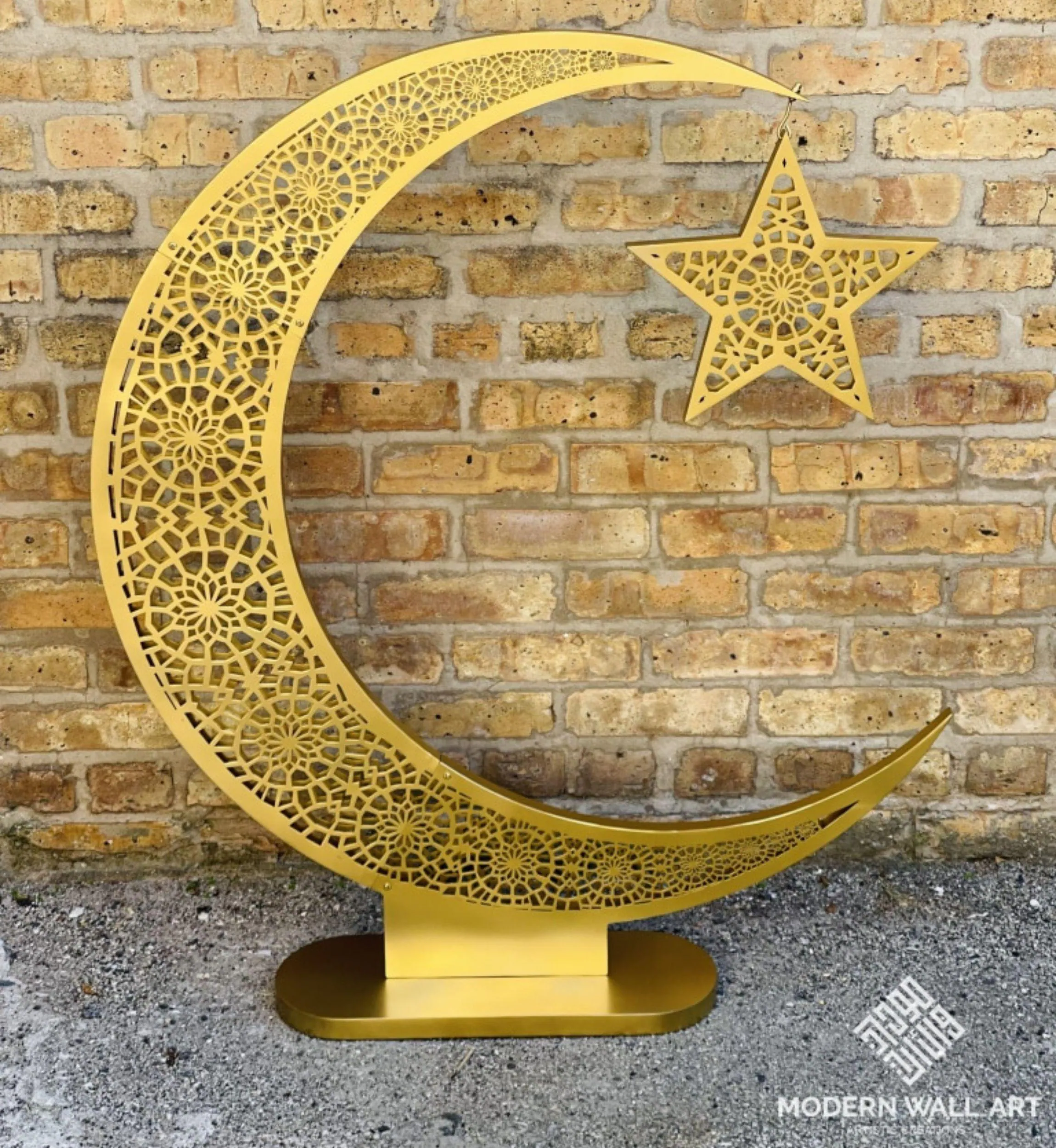 A large, freestanding golden crescent moon with intricate cut-out patterns, holding a matching star by a chain, stands on a flat base against a brick wall.