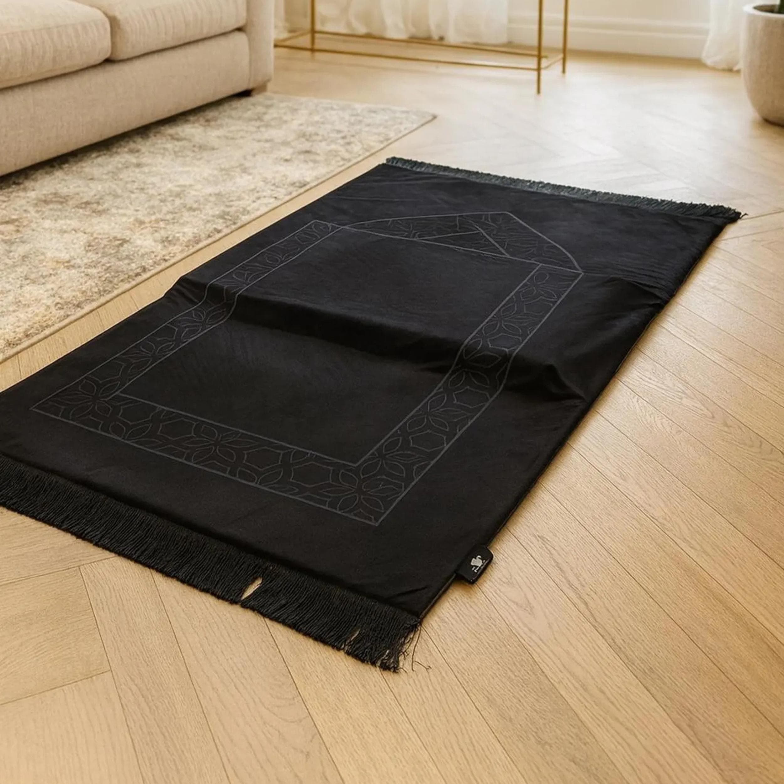 A black prayer mat with a subtle gray geometric pattern and fringed ends lies flat on a light wood floor in a room with a beige sofa and rug.