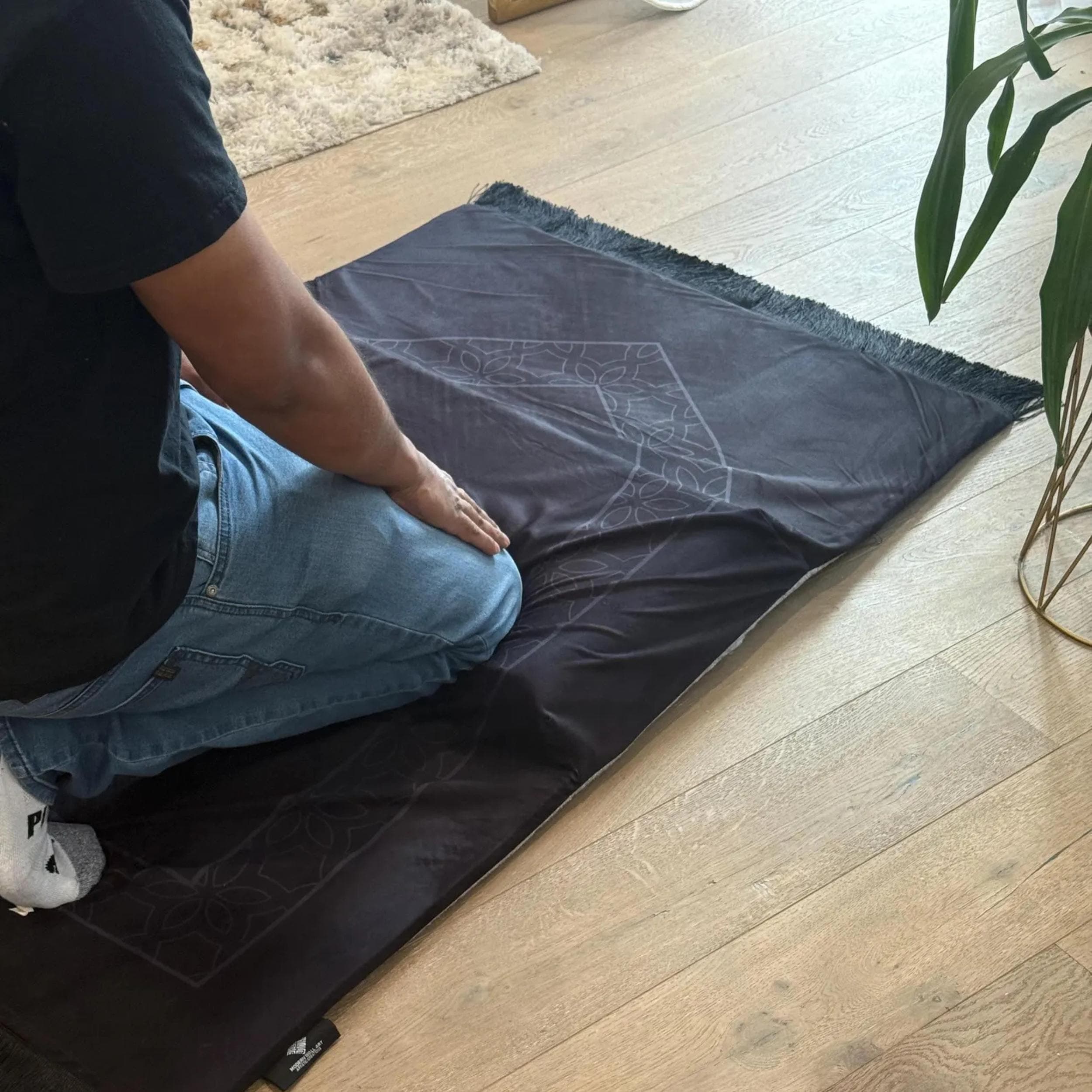 A person kneels on a black prayer mat with a subtle gray geometric pattern, facing away from the camera, with a white sock visible on their left foot.