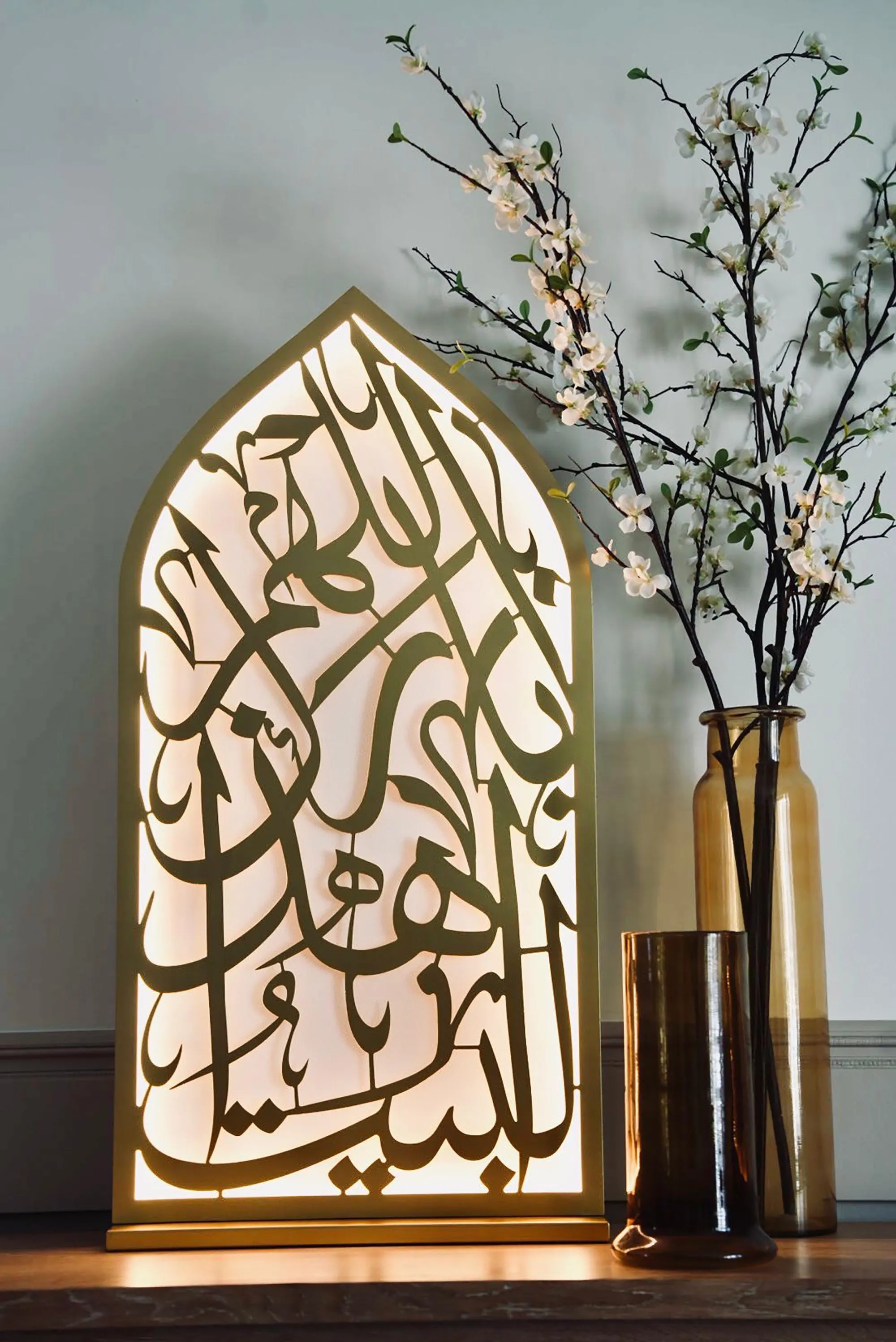 The illuminated mihrab light with gold Arabic calligraphy is positioned on a dark surface, with a vase of white flowering branches and a small amber glass nearby.