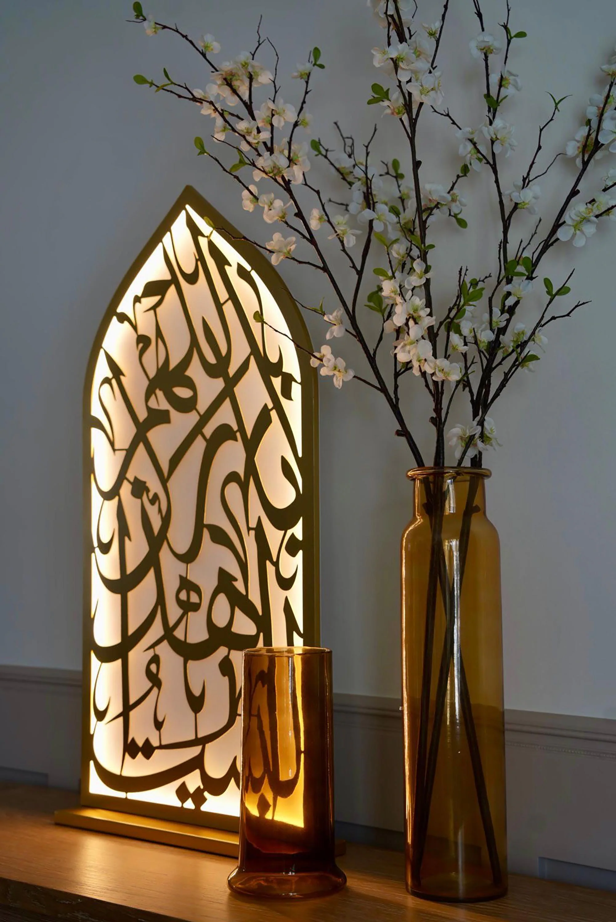 An illuminated mihrab light with gold Arabic calligraphy stands next to a tall amber vase with white flowering branches and a small amber candle holder.