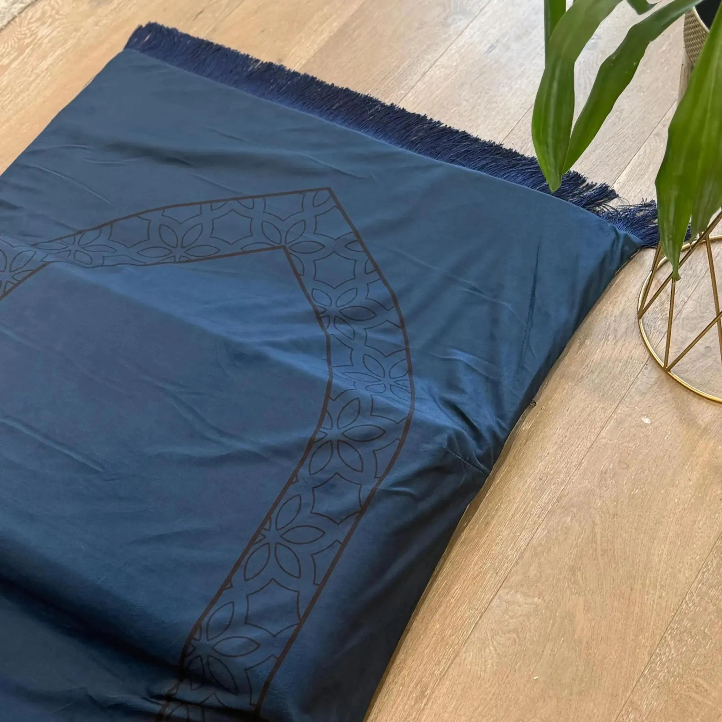 A dark blue prayer mat with a brown geometric pattern and fringed ends is partially rolled up on a light wood floor next to a plant in a gold stand.