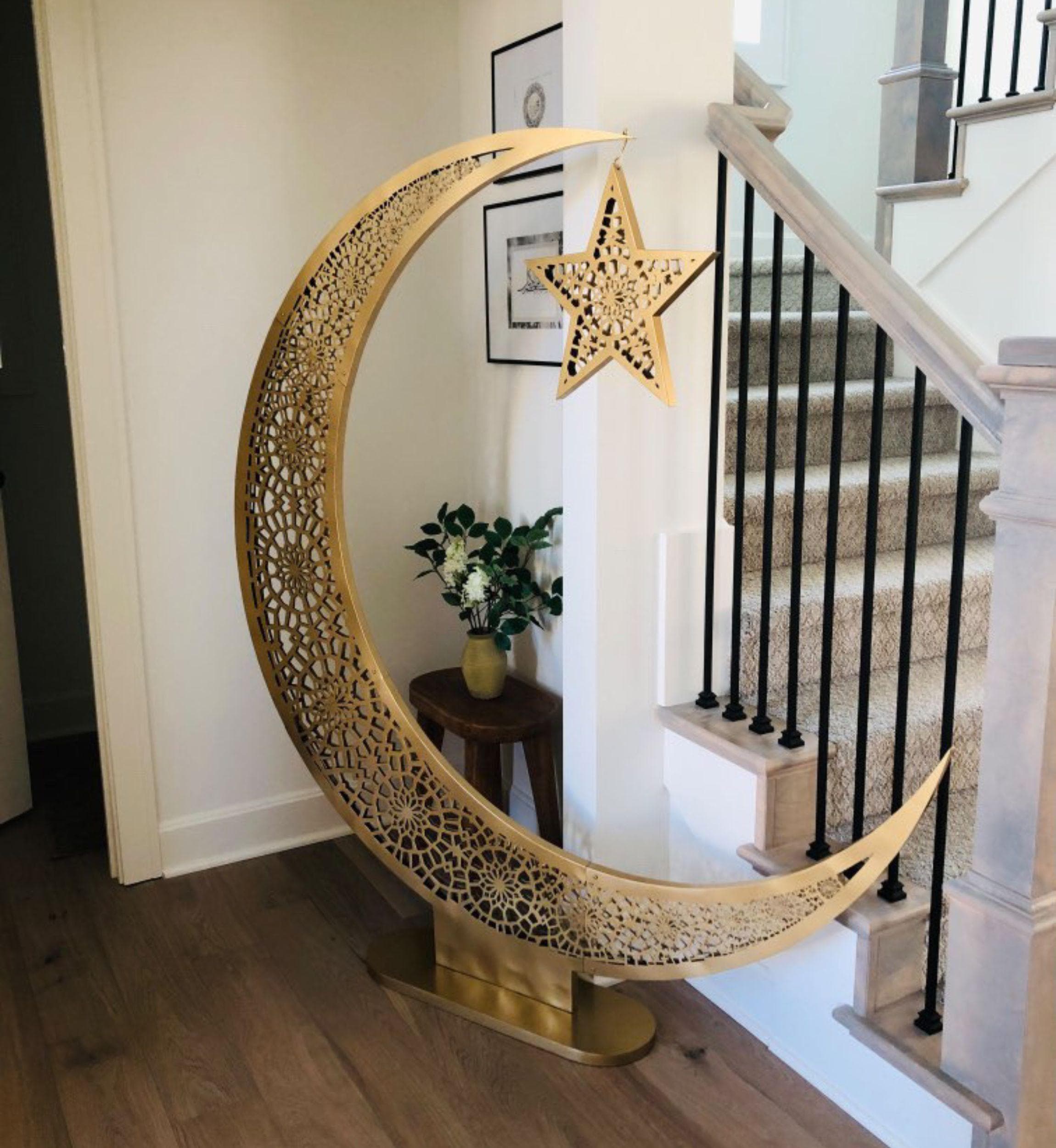 A large, freestanding golden crescent moon with intricate cut-out patterns, holding a matching star by a chain, stands on a flat base next to a staircase.