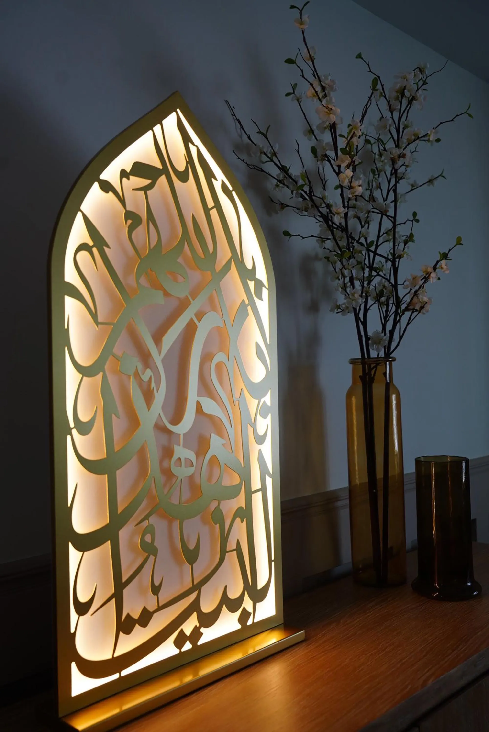 A close-up of the illuminated mihrab light with gold Arabic calligraphy, showing the warm glow behind the metalwork, next to a vase with white flowers.
