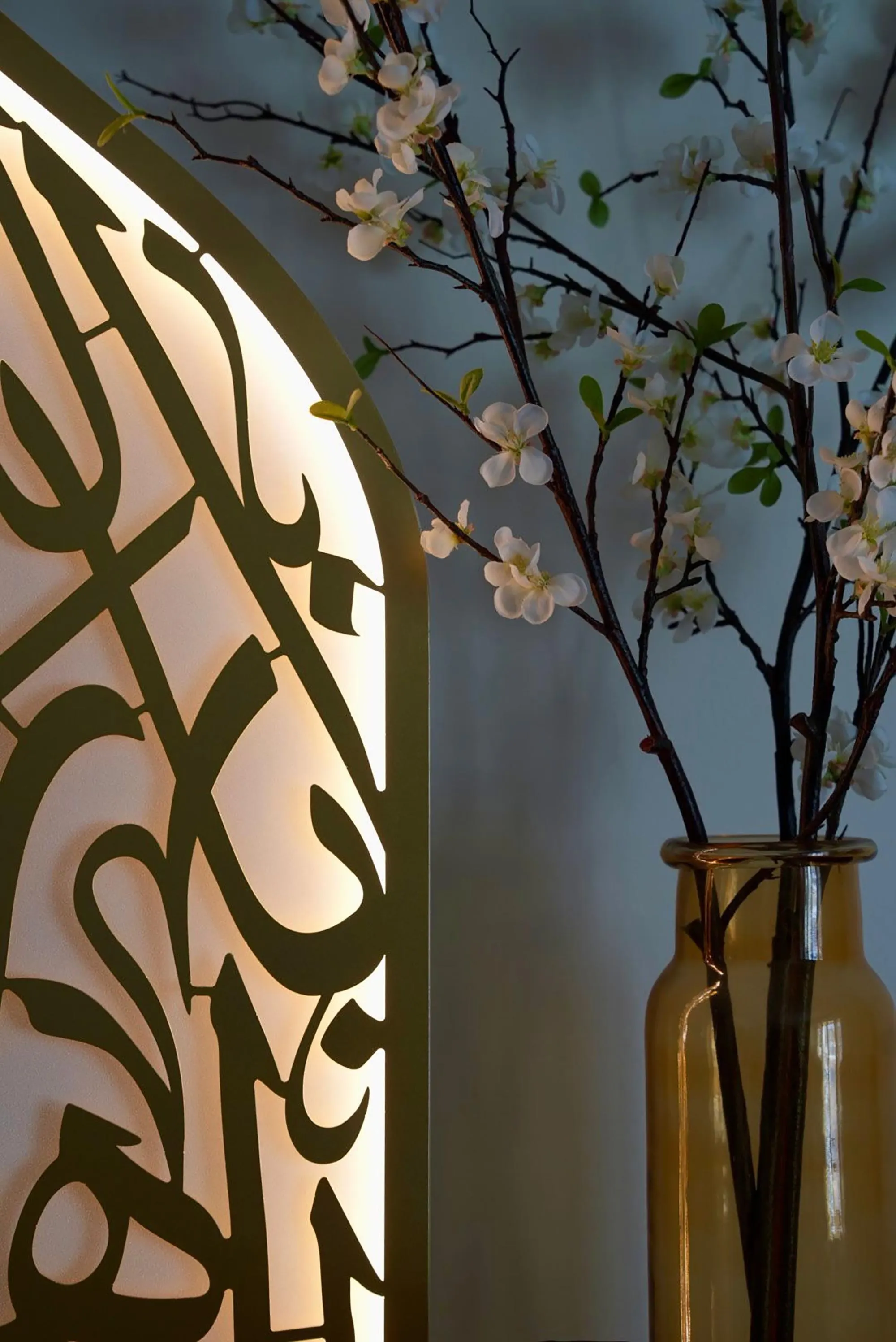 A close-up of the left side of the illuminated mihrab light, showing the gold Arabic calligraphy and the light, next to a vase with white flowering branches.
