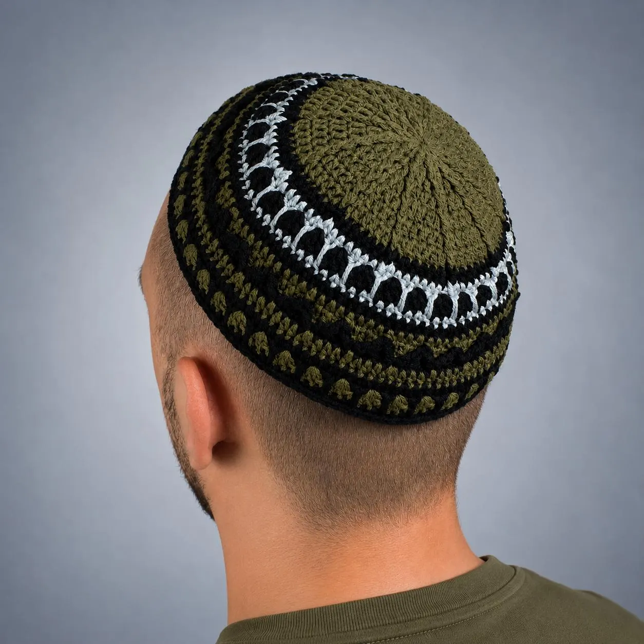 A crocheted kippah in olive green, black, and white. The kippah features a detailed pattern with a circular design. The person wearing the kippah is seen from the side, with a white shirt and a neutral background.