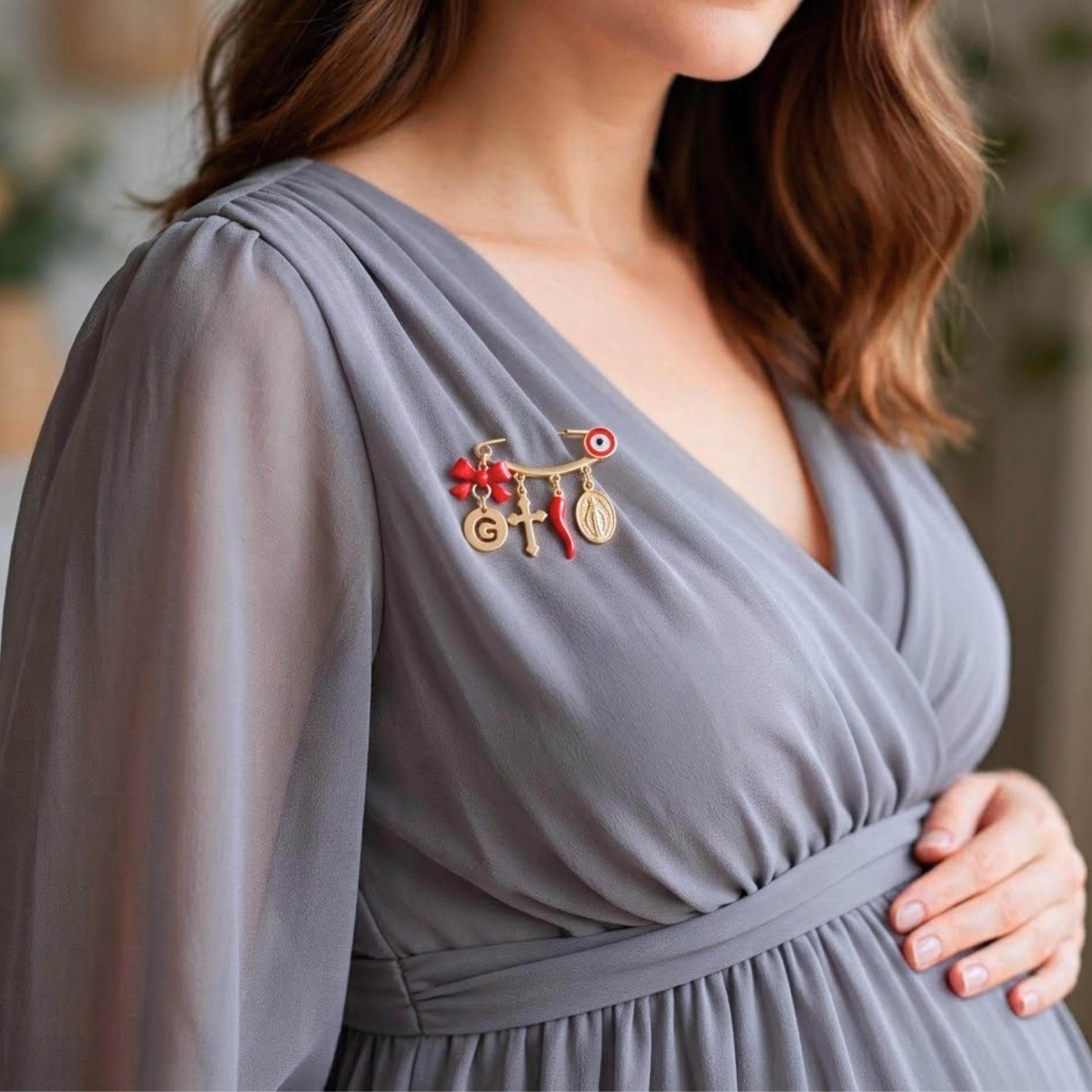 Gold pregnancy safety pin protection brooch featuring initial G disc, red bow, cross, lucky Italian horn cornicello, Virgin Mary Miraculous medal, and evil eye charms pinned to a grey chiffon maternity dress.