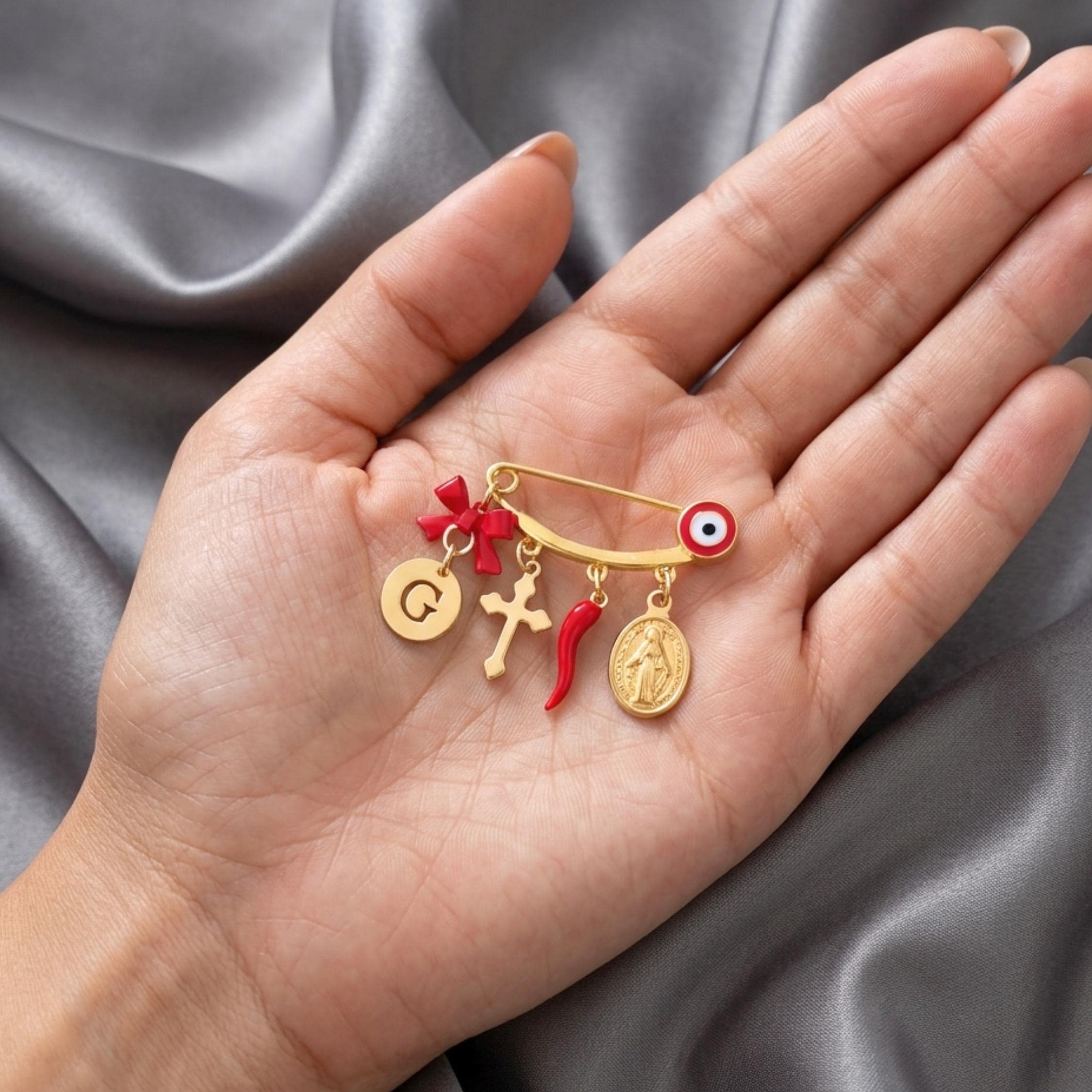 Gold baby safety pin brooch with a red evil eye accent and dangling charms: initial G coin, red bow, cross, red Italian cornicello horn, and Virgin Mary medal. The pin placed on a palm hand.