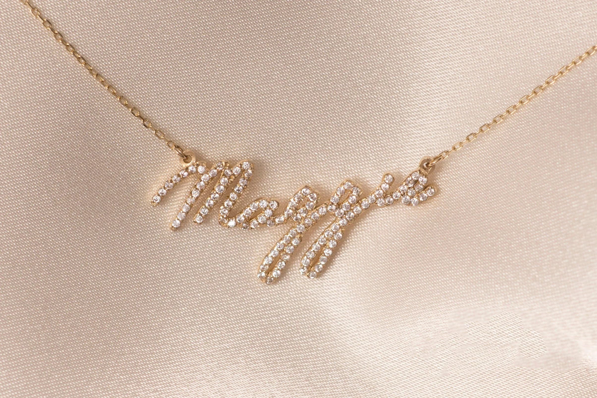 May include: A gold-colored necklace with the name "Maggie" spelled out in cursive script. The letters are encrusted with small, sparkling crystals. The necklace rests on a cream-colored, satin-like fabric.