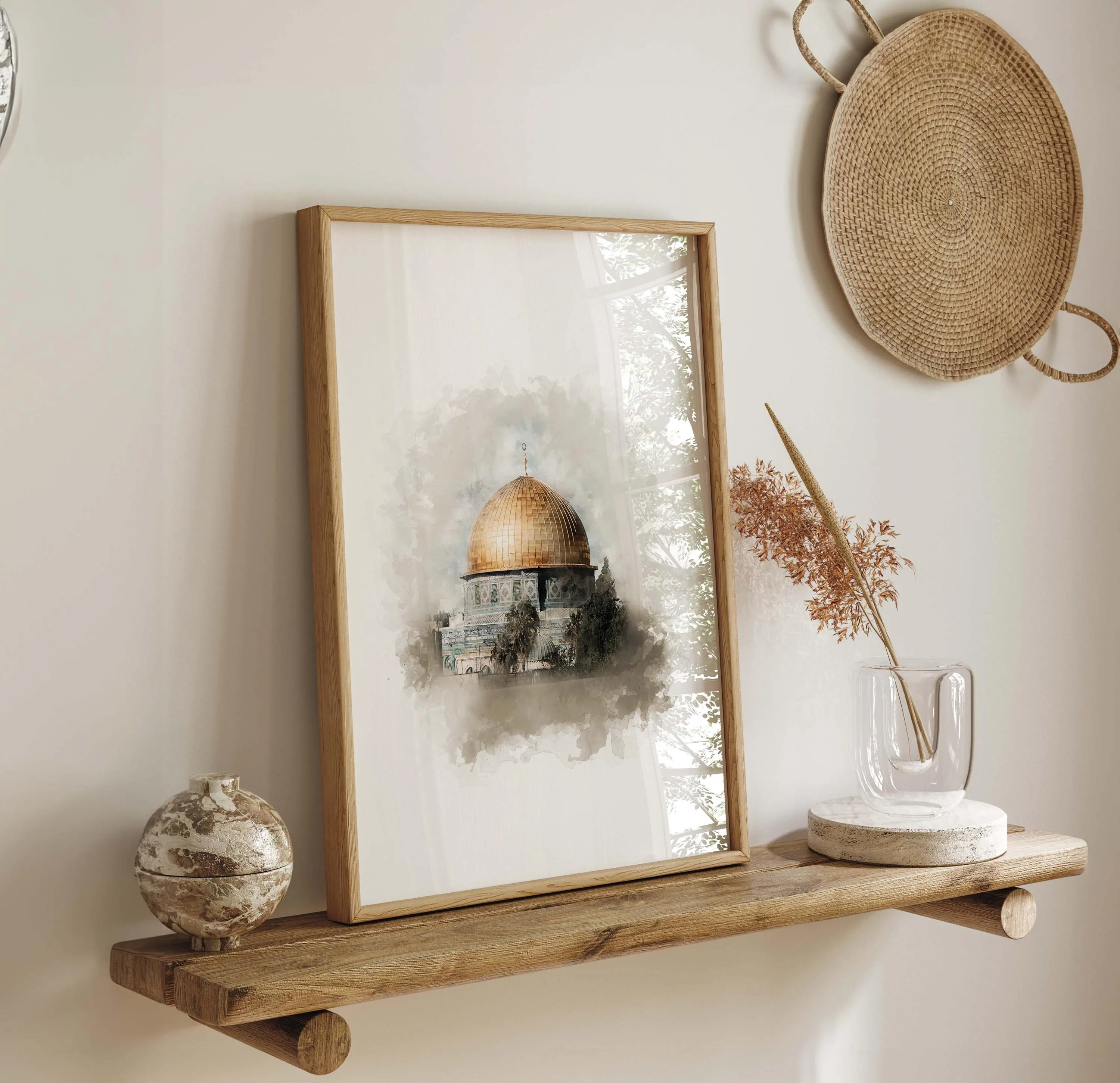 May include: A framed print featuring a watercolor depiction of the Dome of the Rock with a gold dome. The artwork is displayed on a wooden shelf with decorative items, including a small vase and a woven wall hanging. The frame is a light brown color.