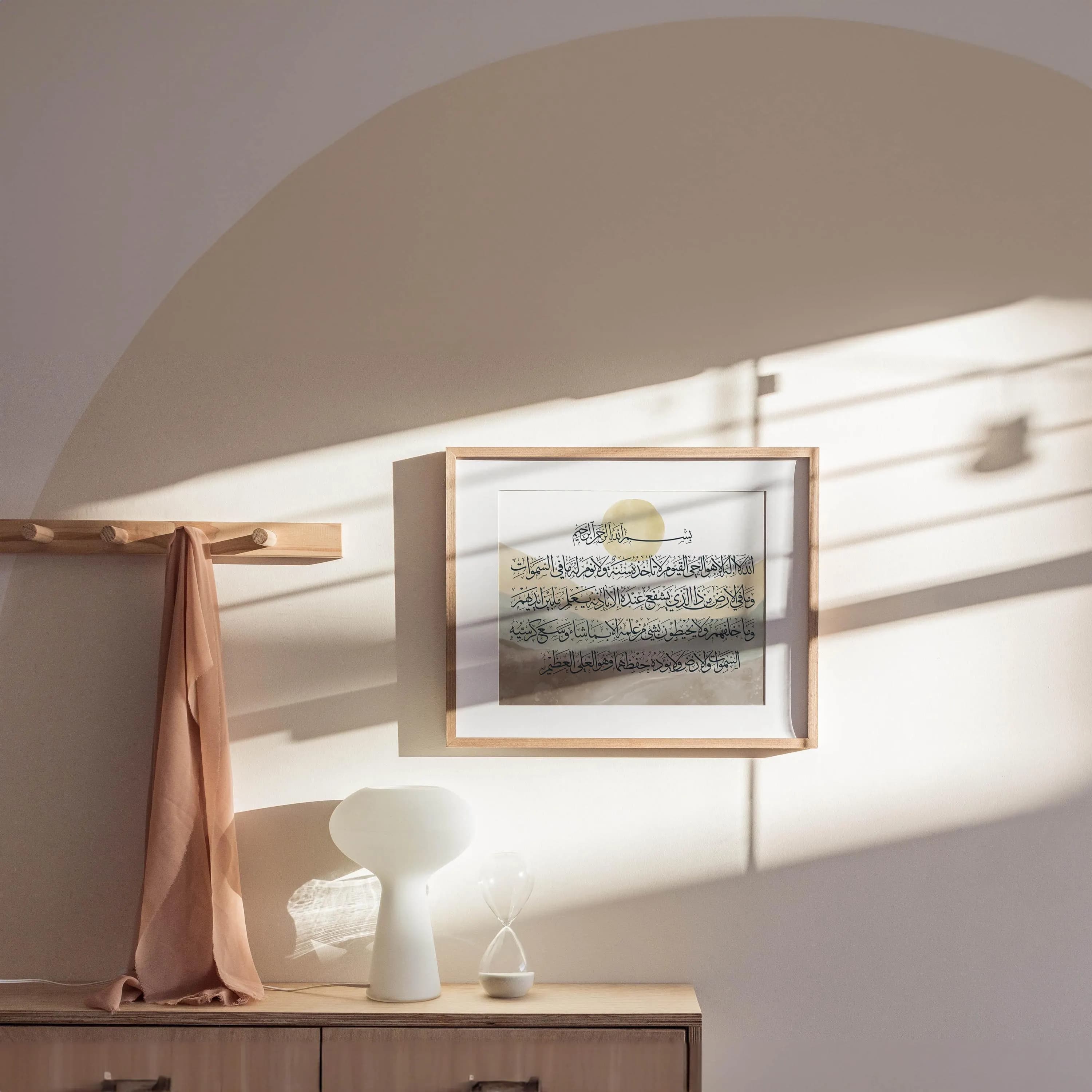 Square wooden frame featuring Ayat al-Kursi Arabic calligraphy with watercolor background of earthy mountains and a soft sun, hung on a wall with warm light and styled alongside a lamp and hanging scarf.