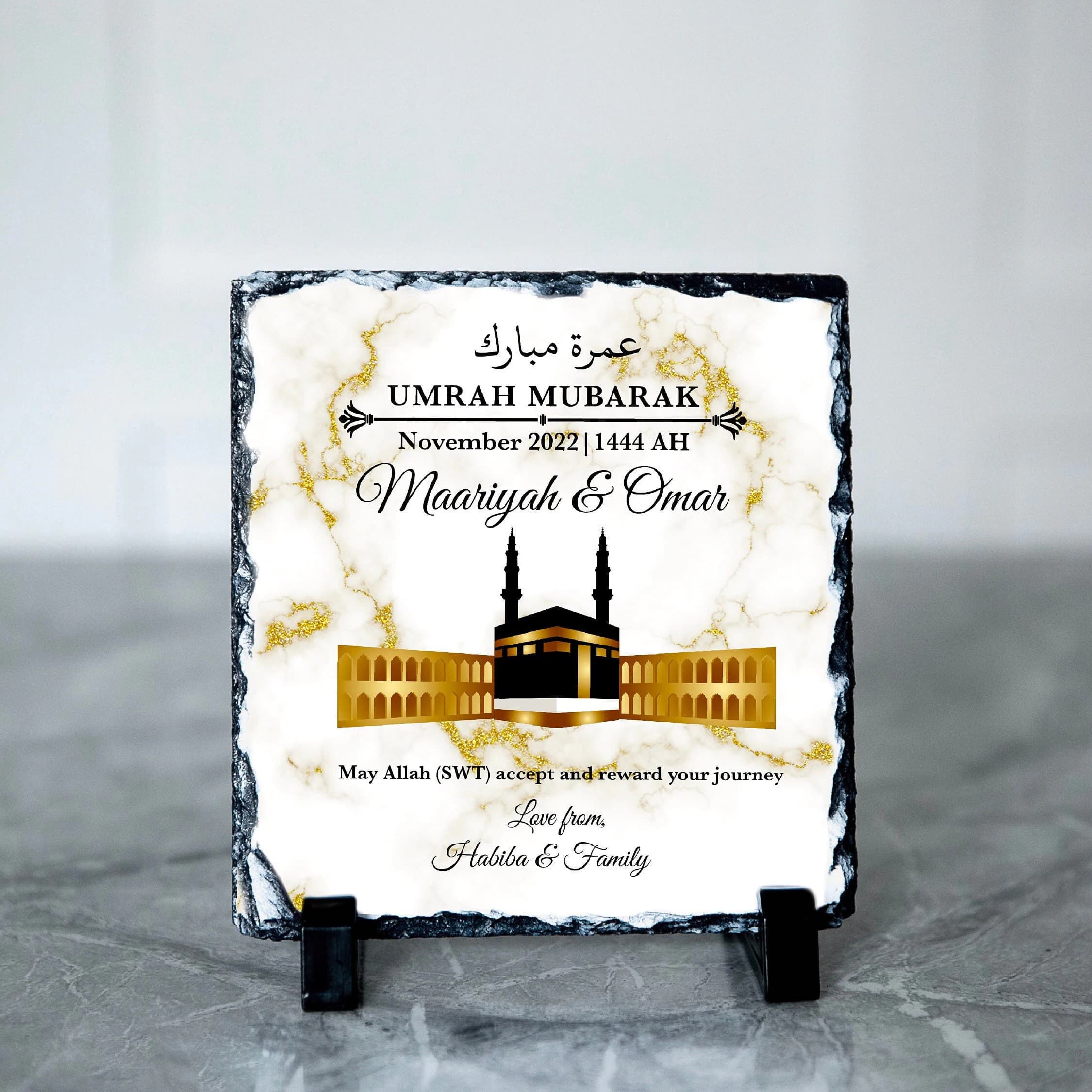 May include: A square slate plaque with a marble effect and gold detailing. The plaque displays the text "UMRAH MUBARAK" and the names "Maariyah & Omar", alongside an illustration of the Kaaba. It is supported by a black stand.