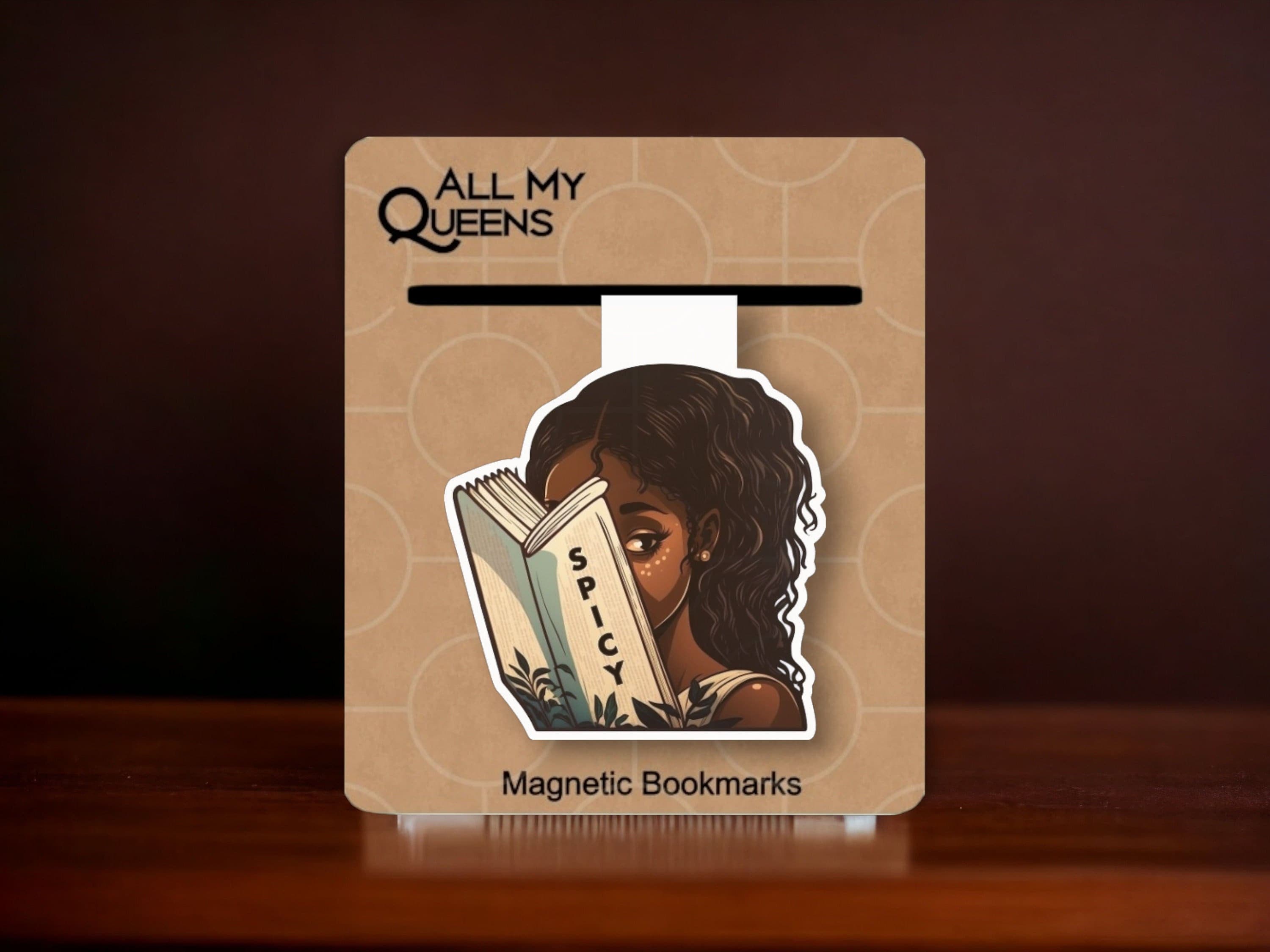 May include: A magnetic bookmark featuring a woman reading a book with the word "Spicy" on the cover. The bookmark is brown and has the text "All My Queens" and "Magnetic Bookmarks" on it.