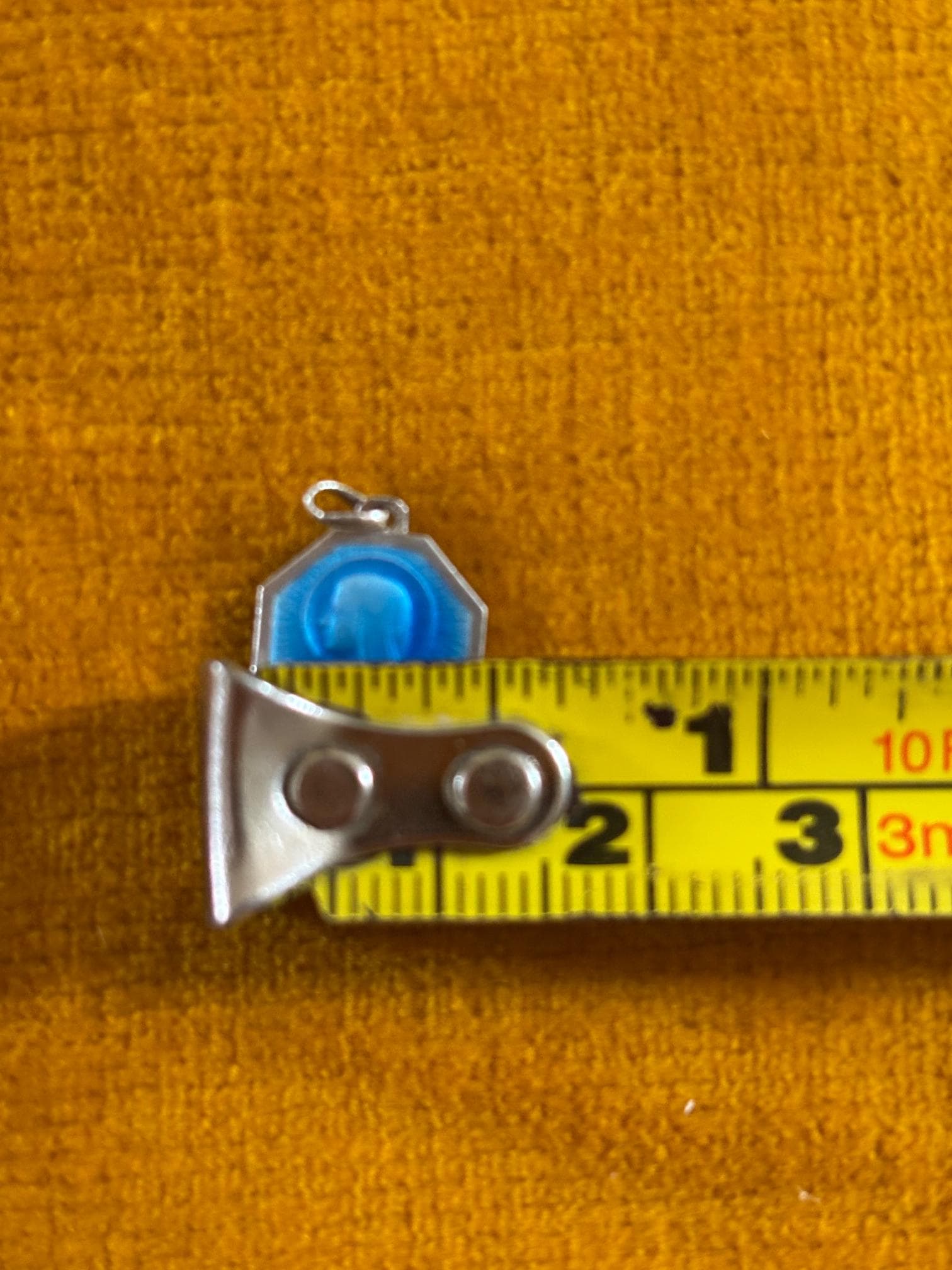 Tiny blue enamel & silver octagonal religious charm shown with tape measure. The charm measures 11mm across
