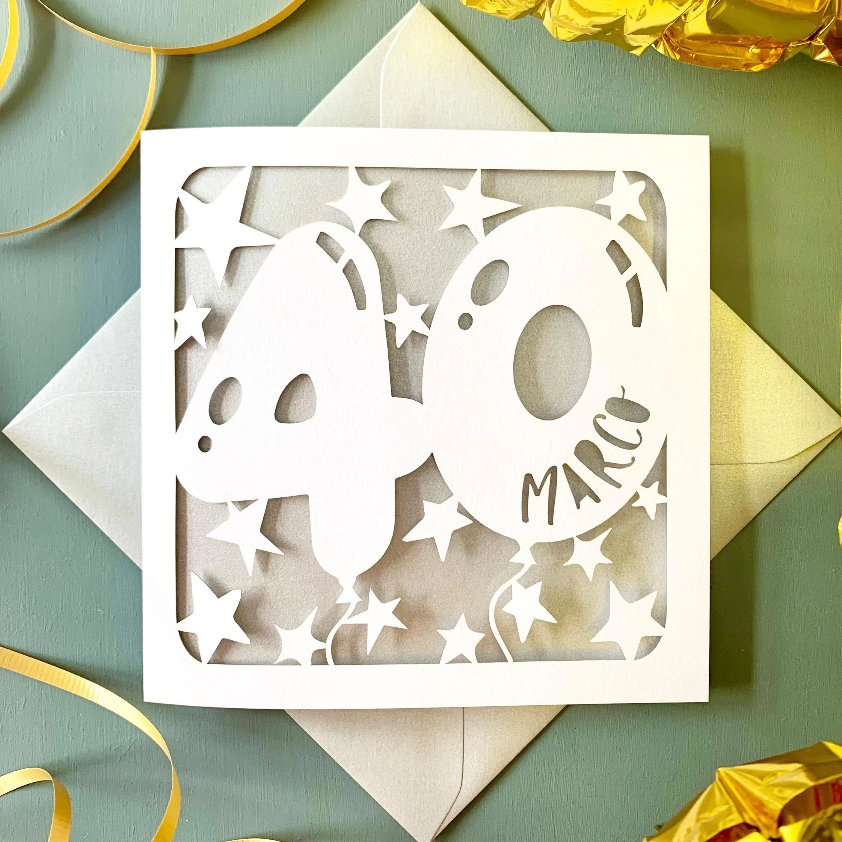 White square laser cut 40th birthday card. Featuring the silhouette of an age 40 balloon, with hand-drawn stars surrounding it. The balloon is personalised with a name in a fun font. The card has a silver paper insert and is lying on a colour-matched envelope. The background is grey with gold decorations visible surrounding.