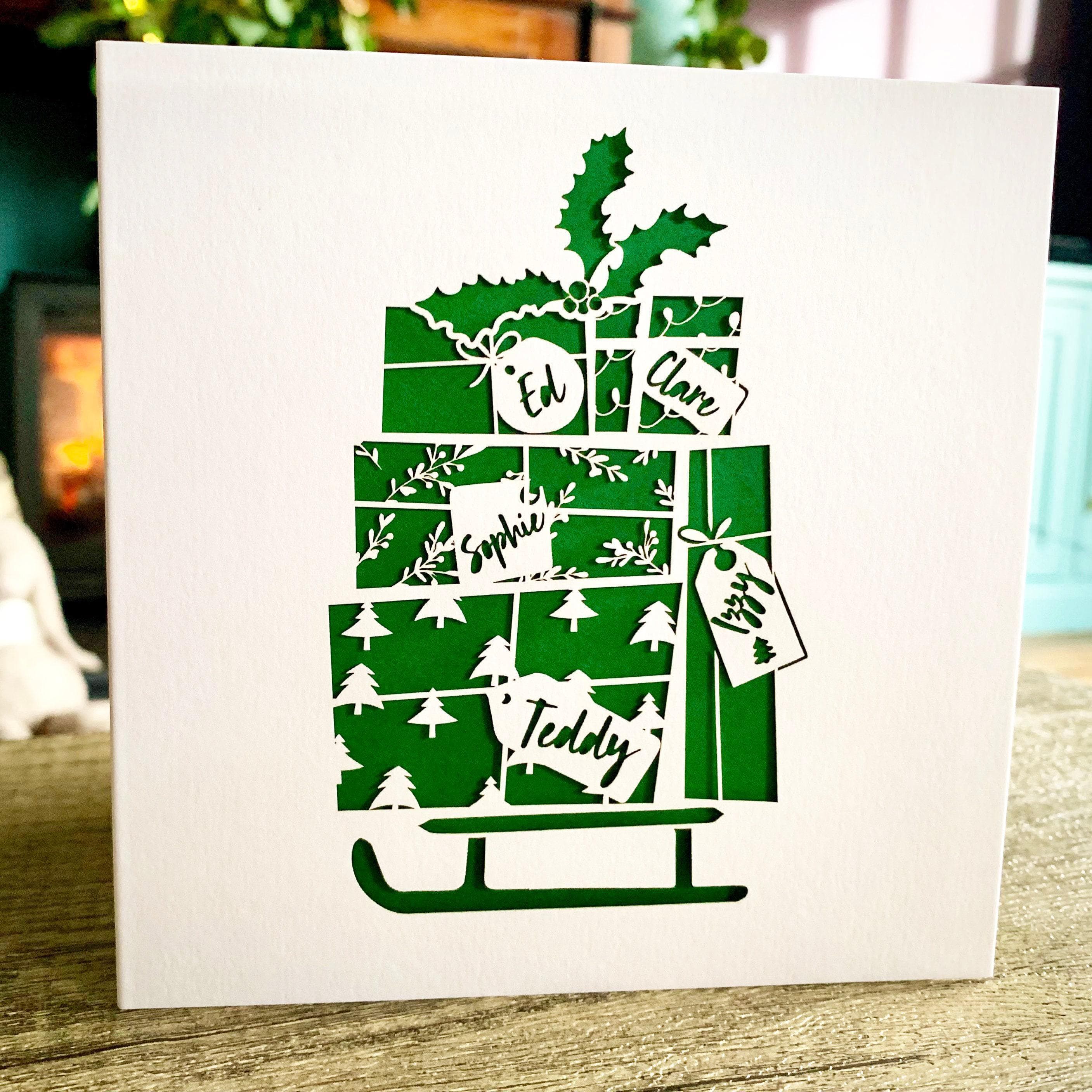 Decorative holiday papercut card featuring a green and white stack of wrapped presents arranged on a sled, topped with holly leaves and berries. Each gift label is personalised with a name. The presents display festive patterns like Christmas trees, snowflakes, and stars. The card is set against a neutral background with a cozy indoor scene softly blurred in the distance.