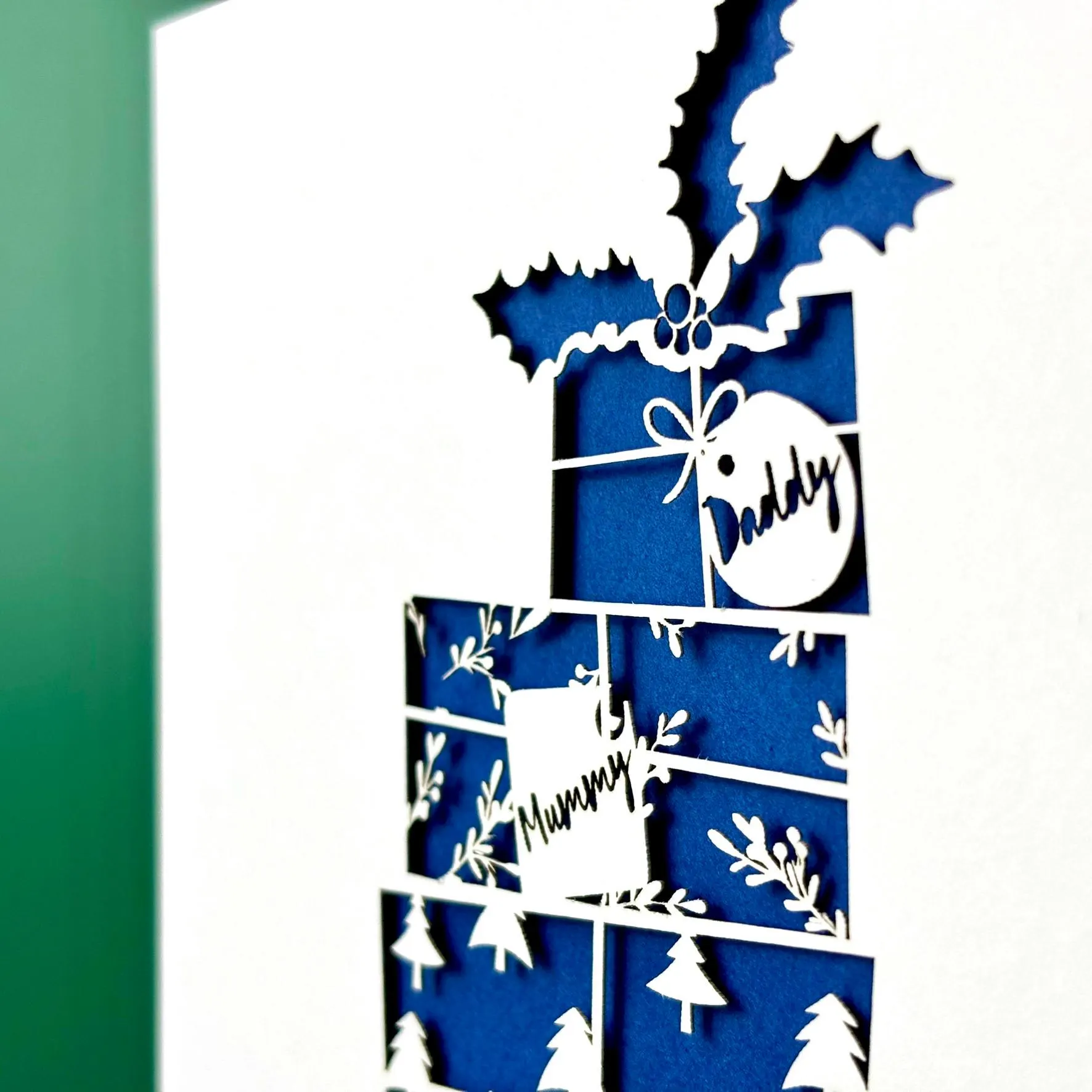Close up of a festive Christmas paper cut card featuring a stylized stack of wrapped presents on a sled, with patterns of trees and stars and topped with a holly sprig. Visible gift tags on each present are personalised with names. The card has a blue paper insert, and rests on a blue envelope.