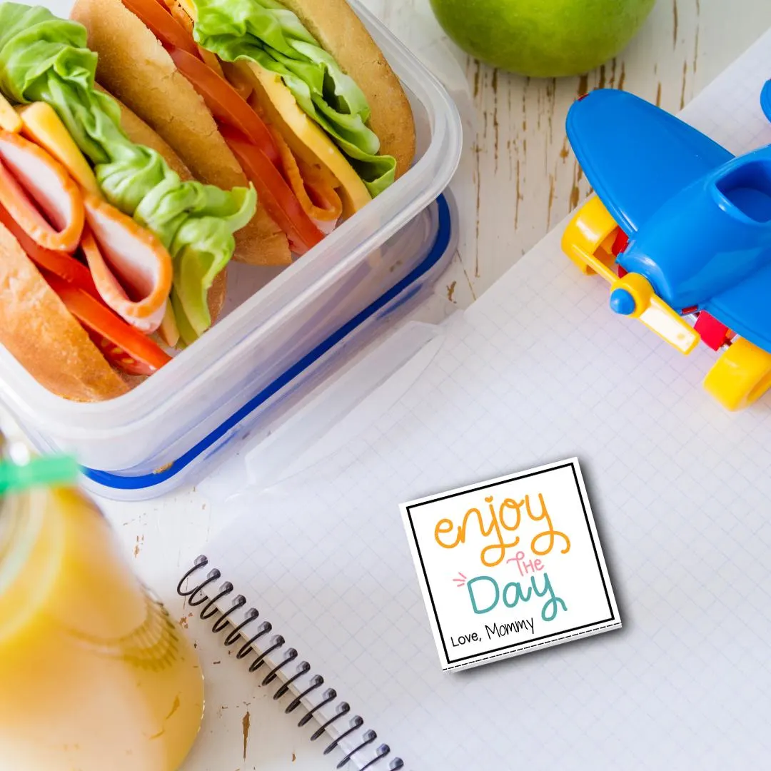 Lunchbox note on top of a notebook signed by mommy with an open lunchbox on the table with a sandwich.