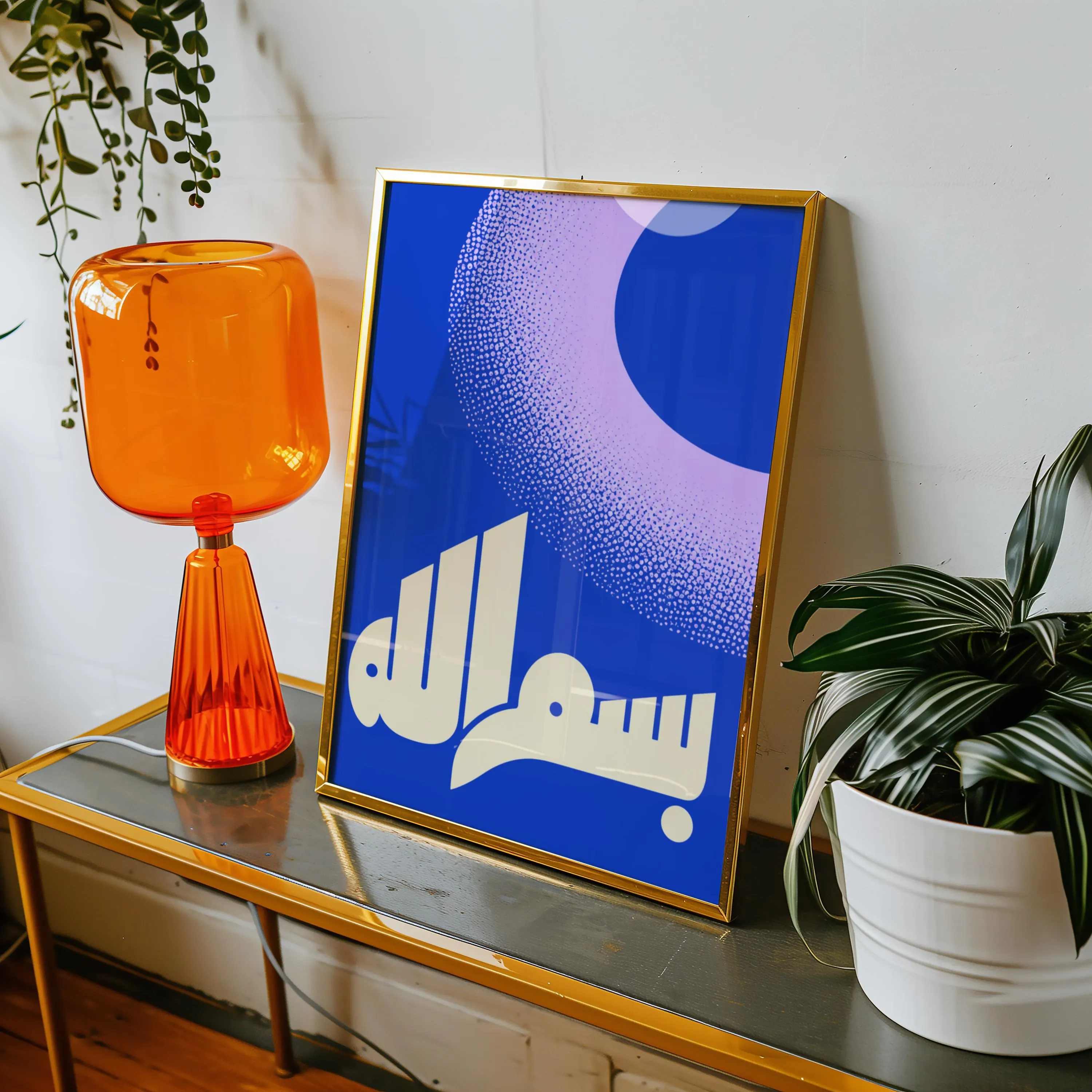 A rectangular gold-framed print featuring a deep blue background with a large, light purple stippled crescent shape and white Arabic calligraphy at the bottom. The artwork is displayed on a surface next to an orange glass lamp.