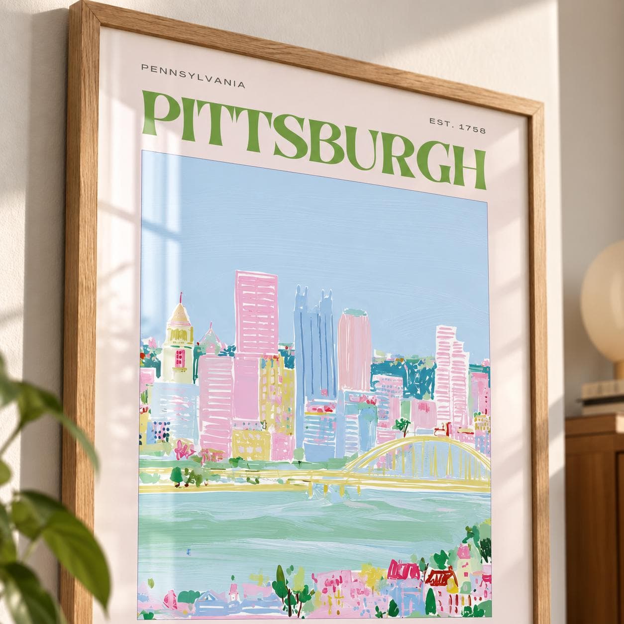 A framed art print of the Pittsburgh skyline featuring the text PENNSYLVANIA and EST. 1758 at the top. The illustration uses a palette of light blue, pink, and green to depict city buildings, a bridge, and a river in a painterly style.