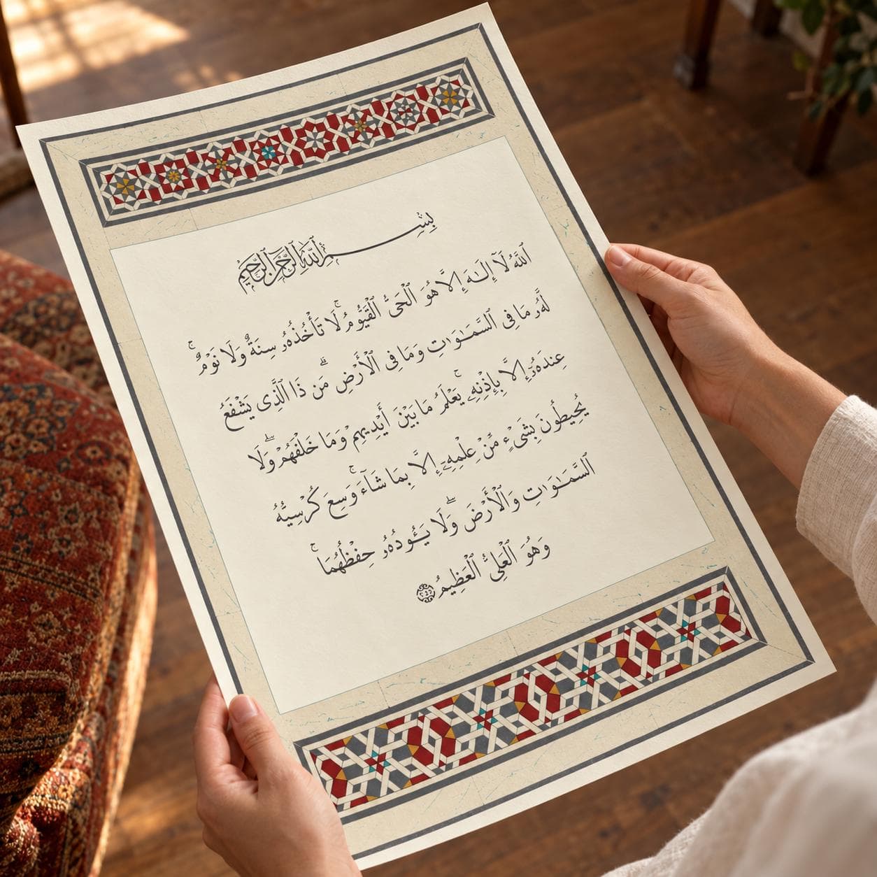 A rectangular paper print featuring black Arabic calligraphy of the Ayat al-Kursi centered on a cream background. The top and bottom borders display a geometric pattern in red, blue, and gold colors on a white base.