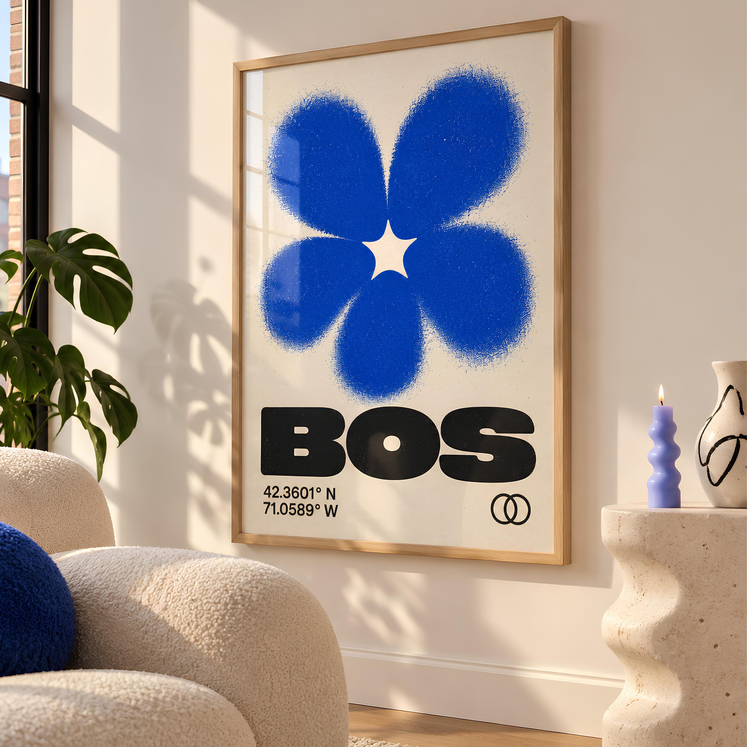 A framed wall art print featuring a large, textured blue flower graphic above the bold black text "BOS". Below the text, the coordinates "42.3601° N 71.0589° W" are printed alongside a small icon of two interlocking circles.