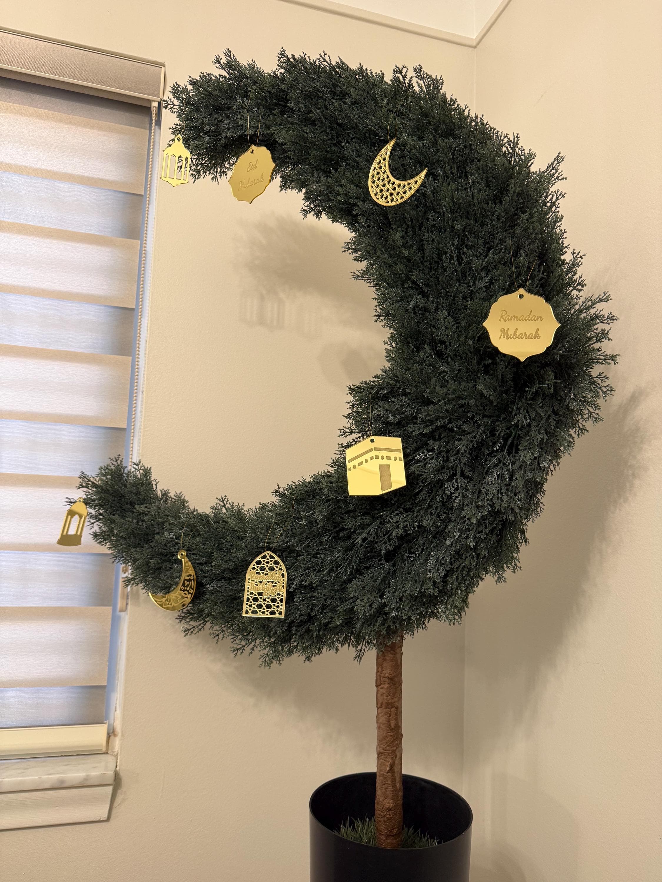 May include: A crescent-shaped decorative tree with dark green foliage and gold-colored ornaments. The ornaments include lanterns, crescent moons, and tags with the text "Ramadan Mubarak". The tree is in a black pot.