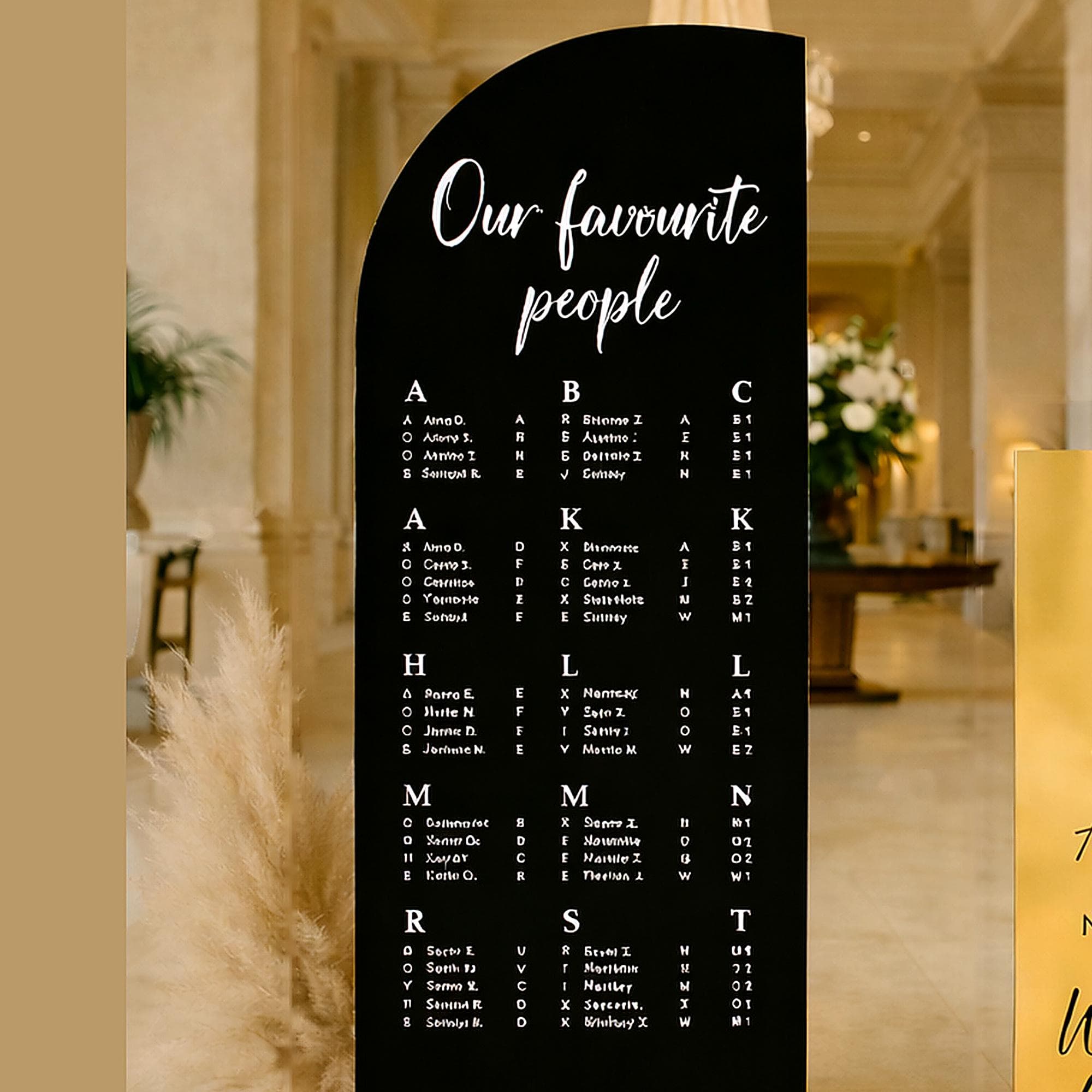 Clearer, less-zoomed close-up of the black acrylic wedding seating chart showing the ‘Our favourite people’ heading and guest list typography, photographed in a warm luxury venue setting