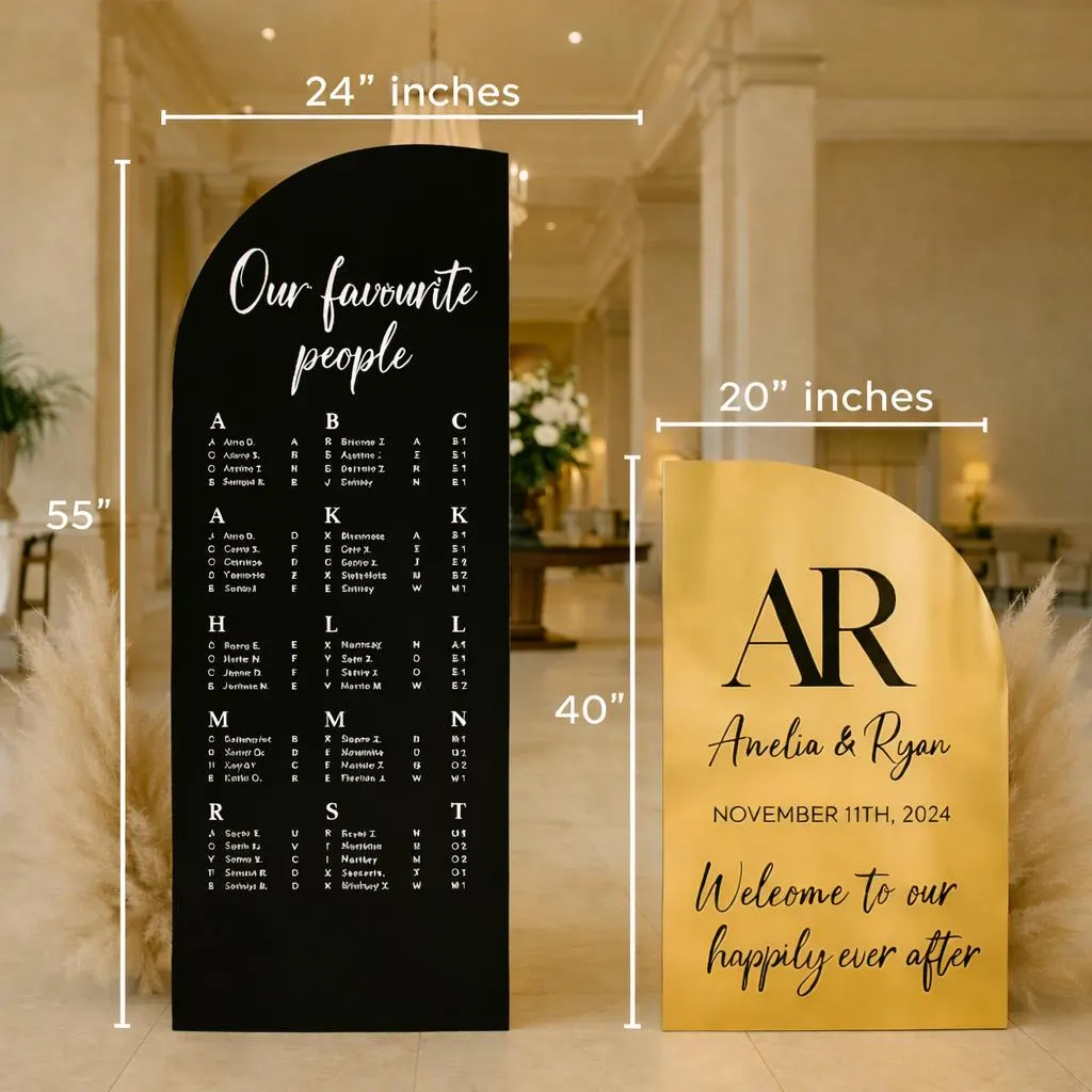 Two freestanding wedding signs in a luxury venue lobby: a tall black seating chart and a gold welcome sign with ‘AR’ monogram, pampas grass accents, and a chandelier overhead