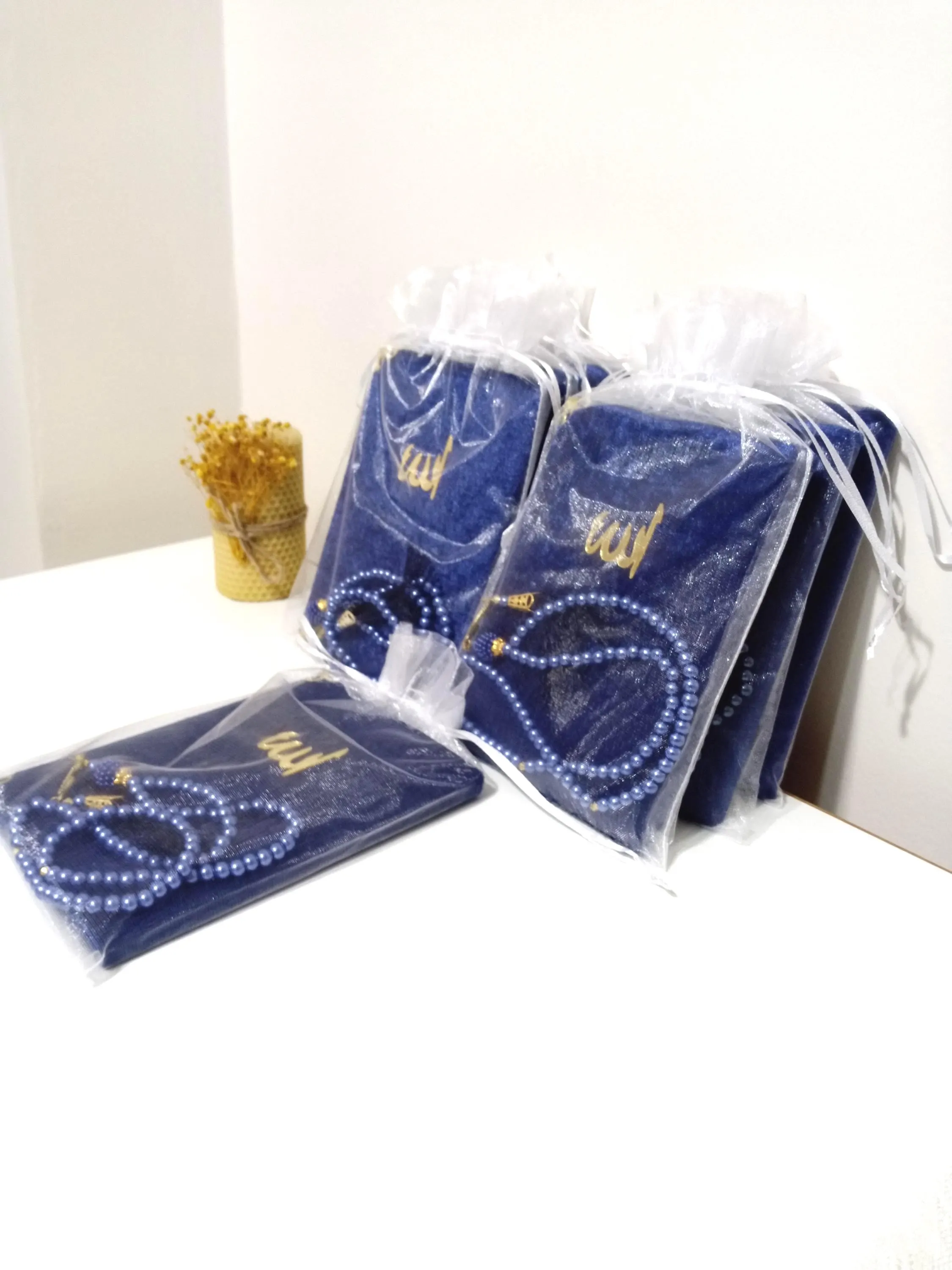May include: A set of blue prayer rugs with gold embroidery and matching blue prayer beads in clear plastic bags. The bags are tied with white ribbon.