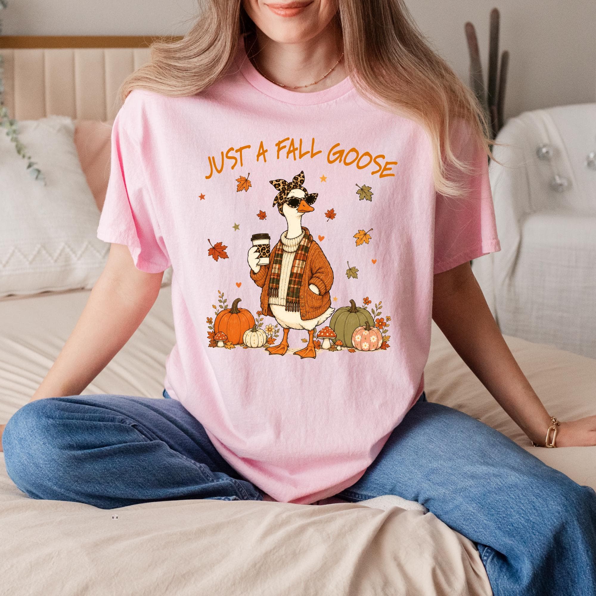 A woman wearing a pink t-shirt with a cartoon goose and autumn decorations, sitting on a bed.