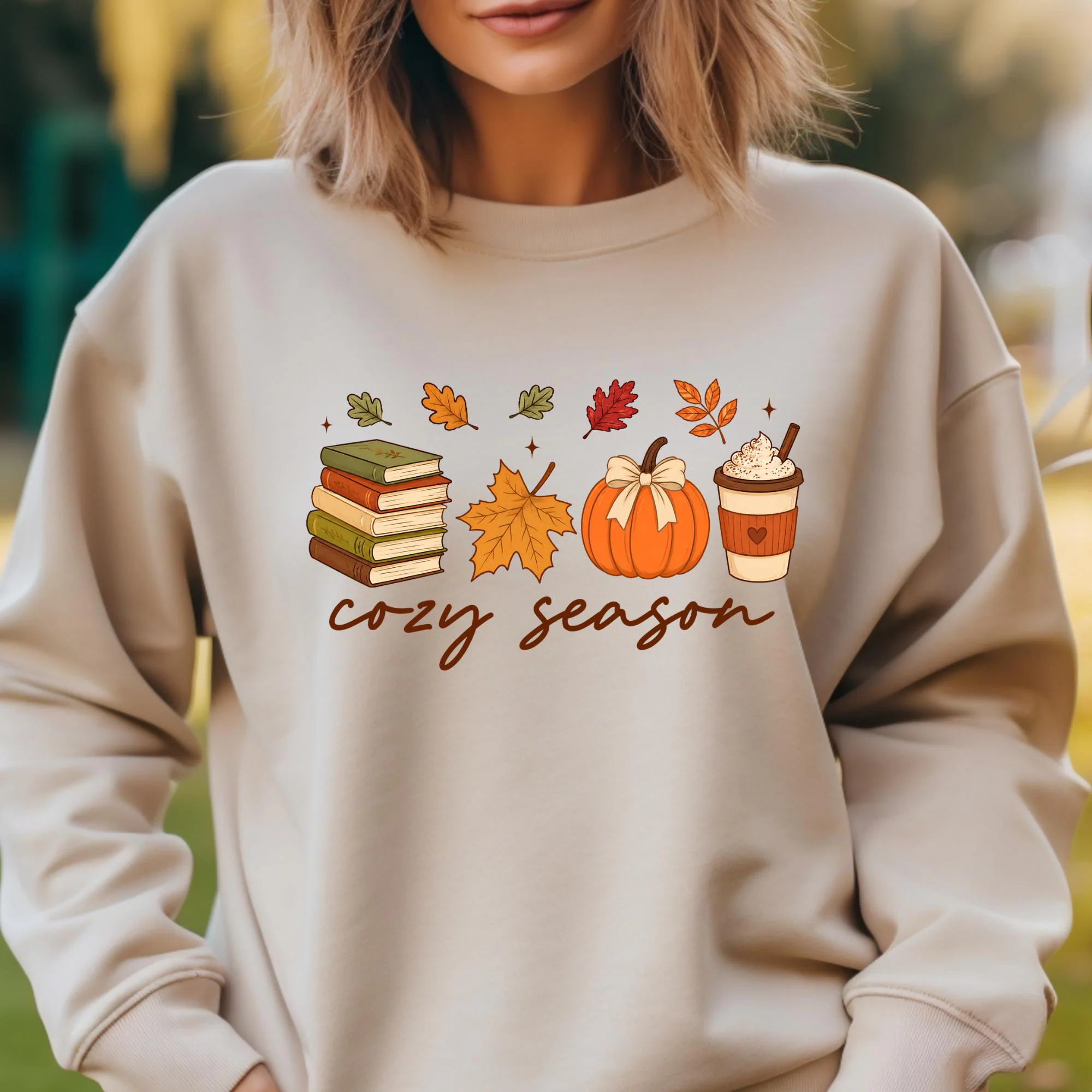 A woman wearing a grey sweatshirt with a graphic design featuring autumn-themed illustrations and the text "cozy season" printed on it.