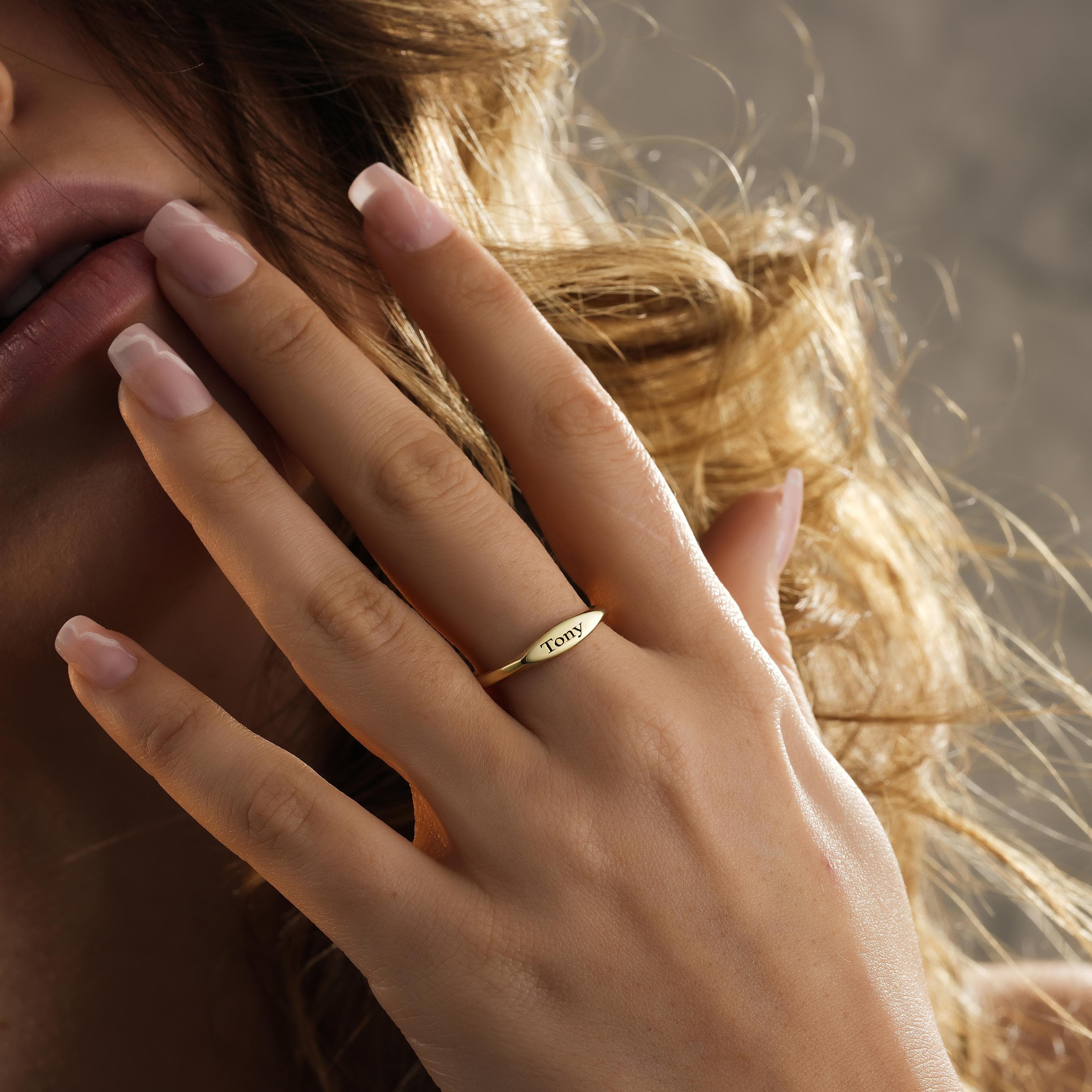 May include: A gold-colored ring with the name "Tony" engraved on a slender, oval-shaped plate. The ring is worn on a finger, showcasing its delicate design and polished finish. The ring's simple elegance makes it a timeless piece of jewelry.