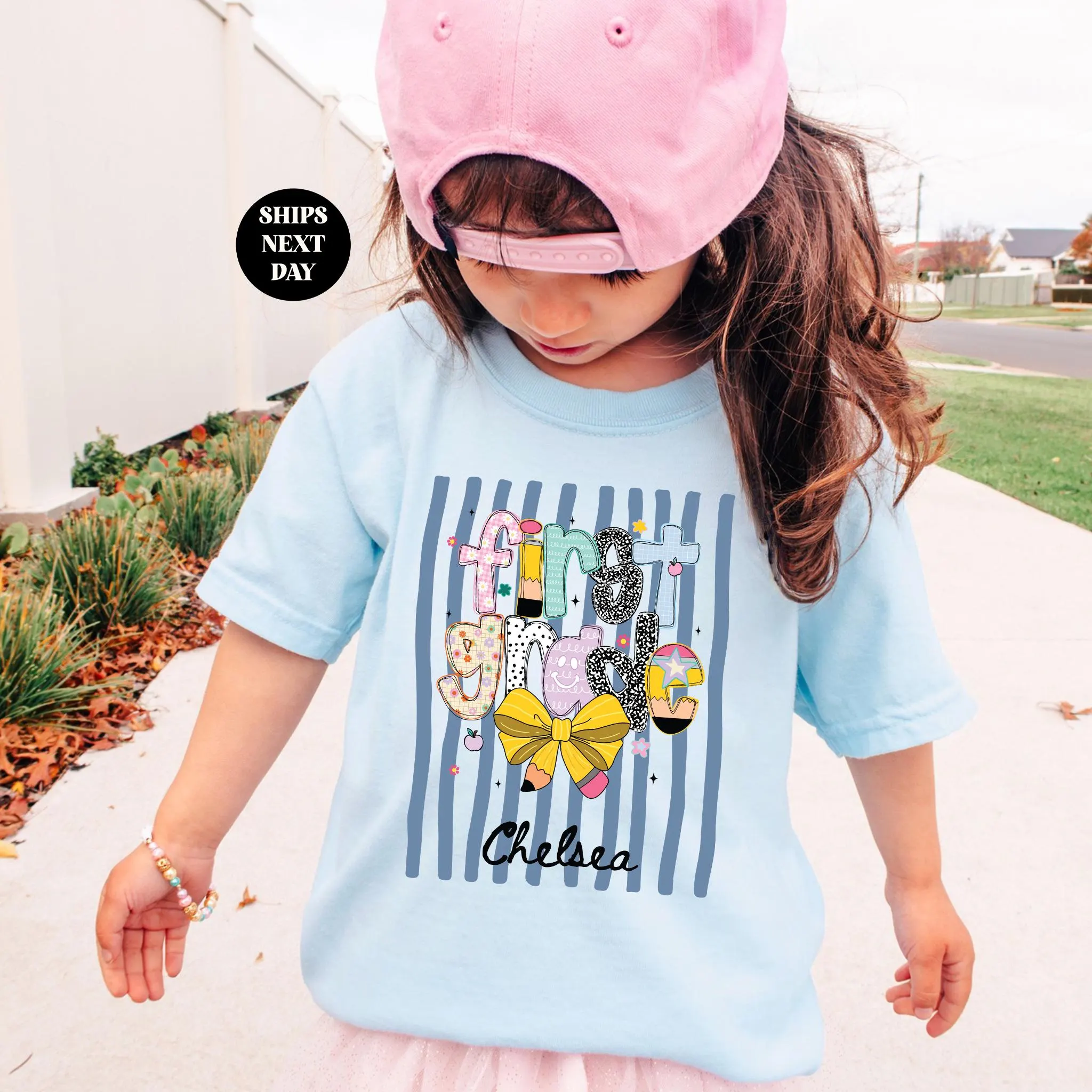 A young girl wearing a pink cap and a light blue t-shirt with a colorful graphic design, standing on a sidewalk.