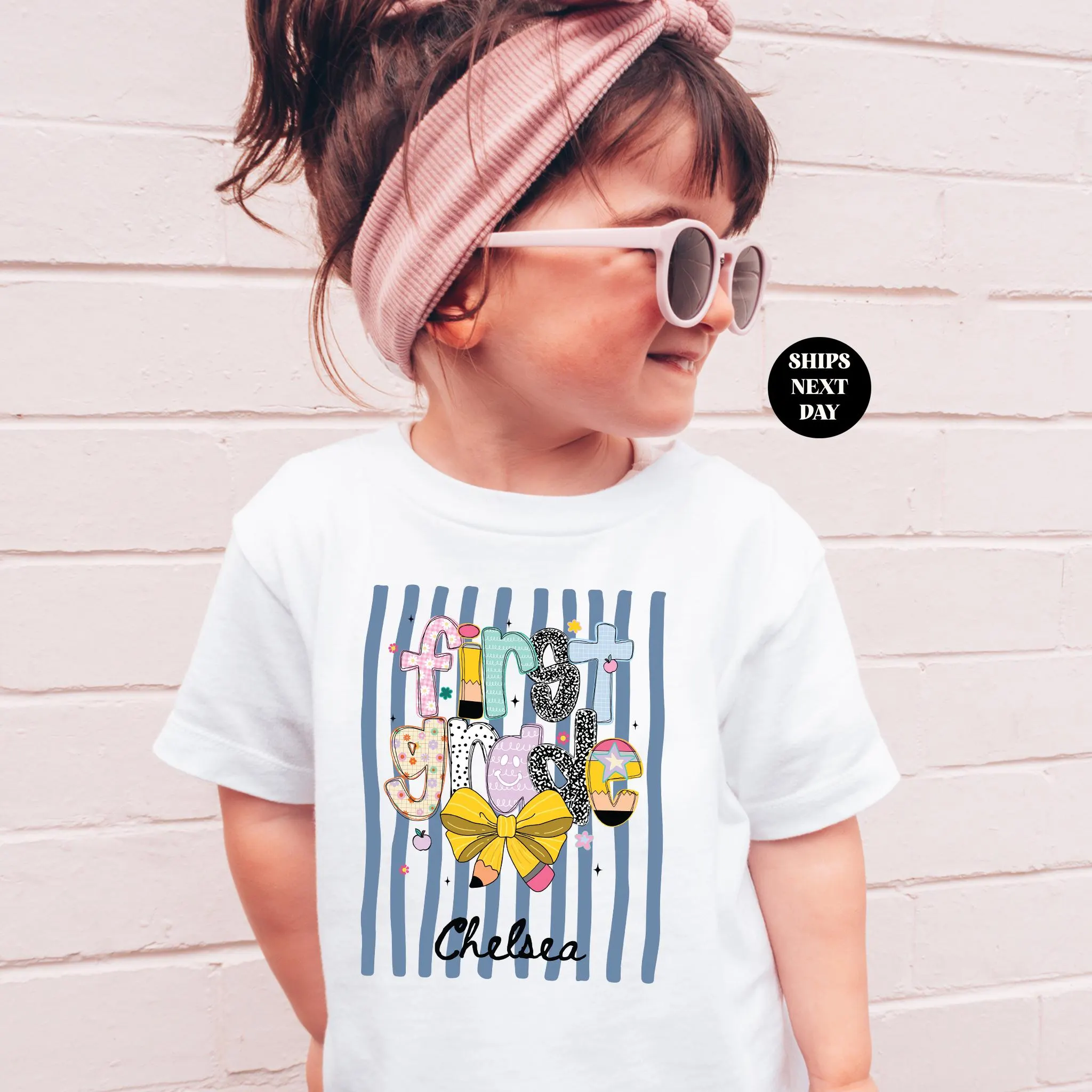 A young girl wearing a white t-shirt with a colorful graphic design and text, standing in front of a white brick wall.