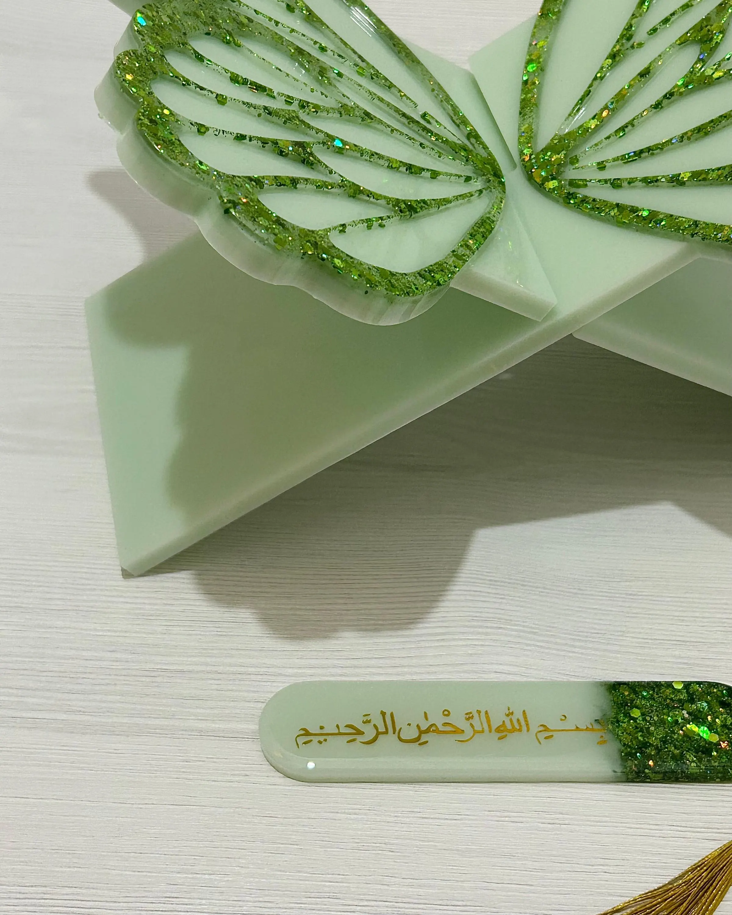 Bottom end of a resin free standing butterfly design Quran stand in sage green and green glitter with a matching bookmark with Bismillah in gold Arabic text on a light wooden background