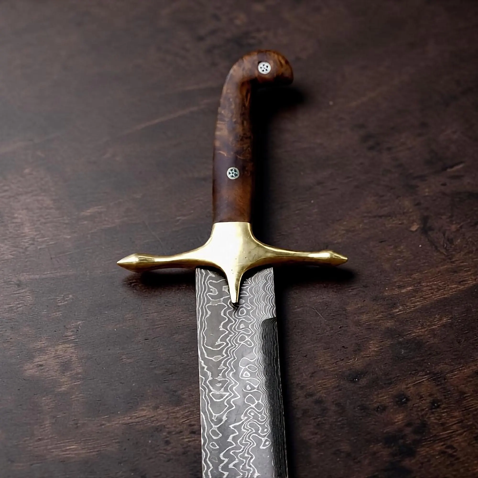 A sword with a patterned Damascus steel blade, a polished brass crossguard, and a curved wooden handle featuring two small circular metal inlays. The blade displays a distinct wavy, layered metal texture throughout its length.