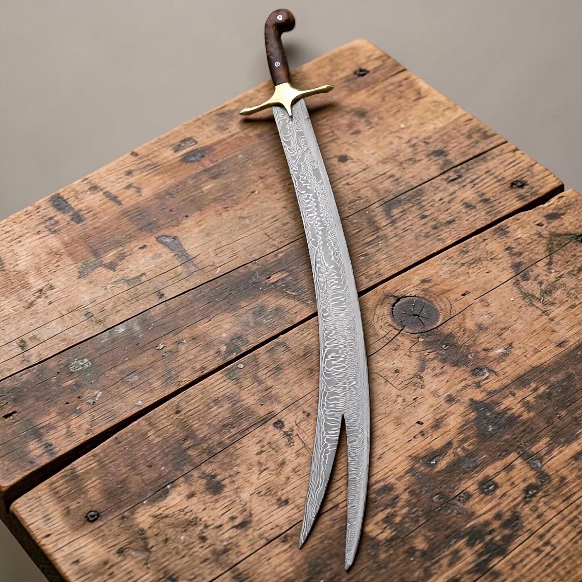 A curved sword with a Damascus steel blade featuring a distinct split tip. The handle is crafted from dark wood with a brass crossguard. The blade displays a complex, wavy pattern throughout its length, ending in two separate points.
