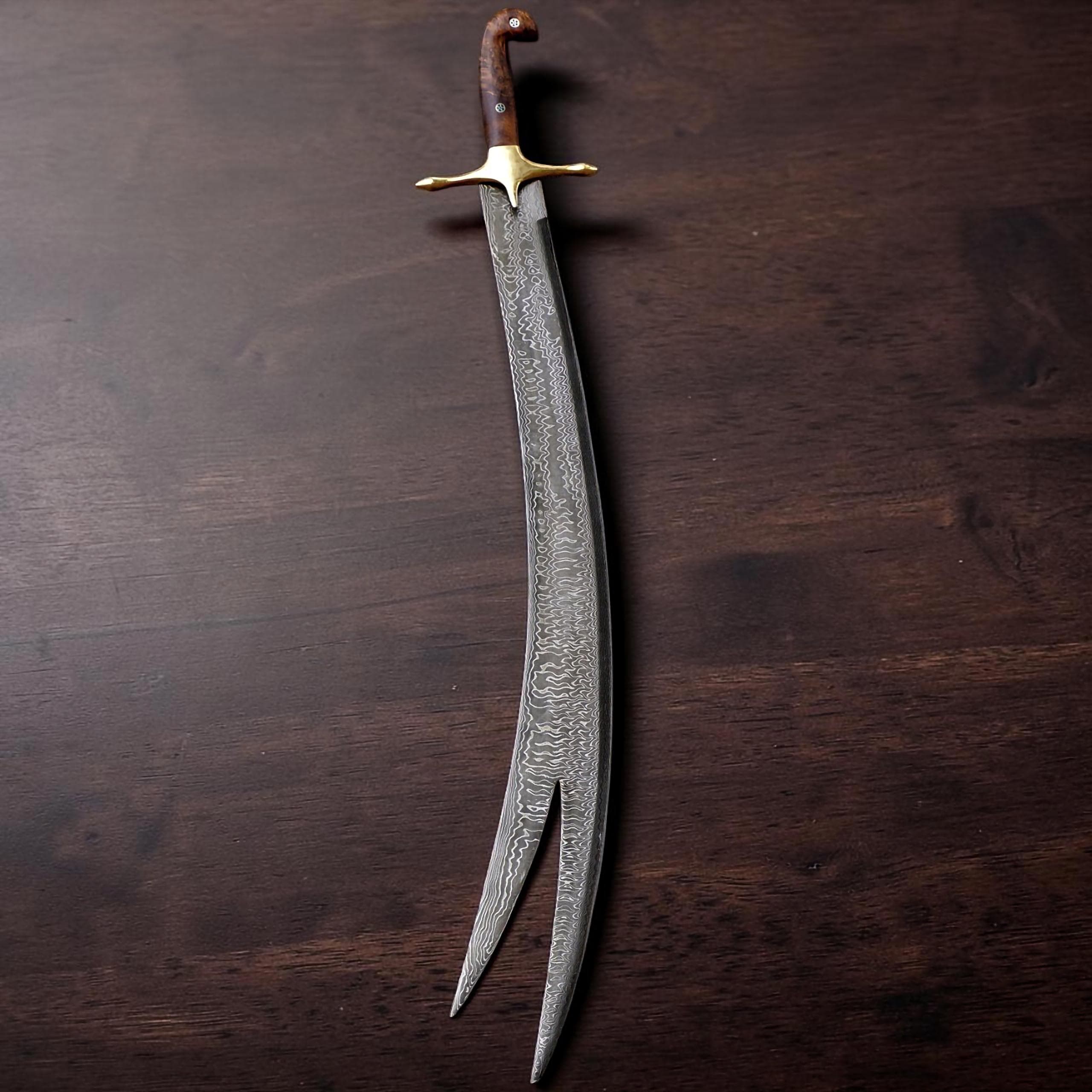 A curved sword featuring a patterned Damascus steel blade with a split tip. The hilt is crafted from dark brown wood with a polished brass crossguard and a single visible rivet, showcasing a distinct, elongated, and bifurcated blade design.