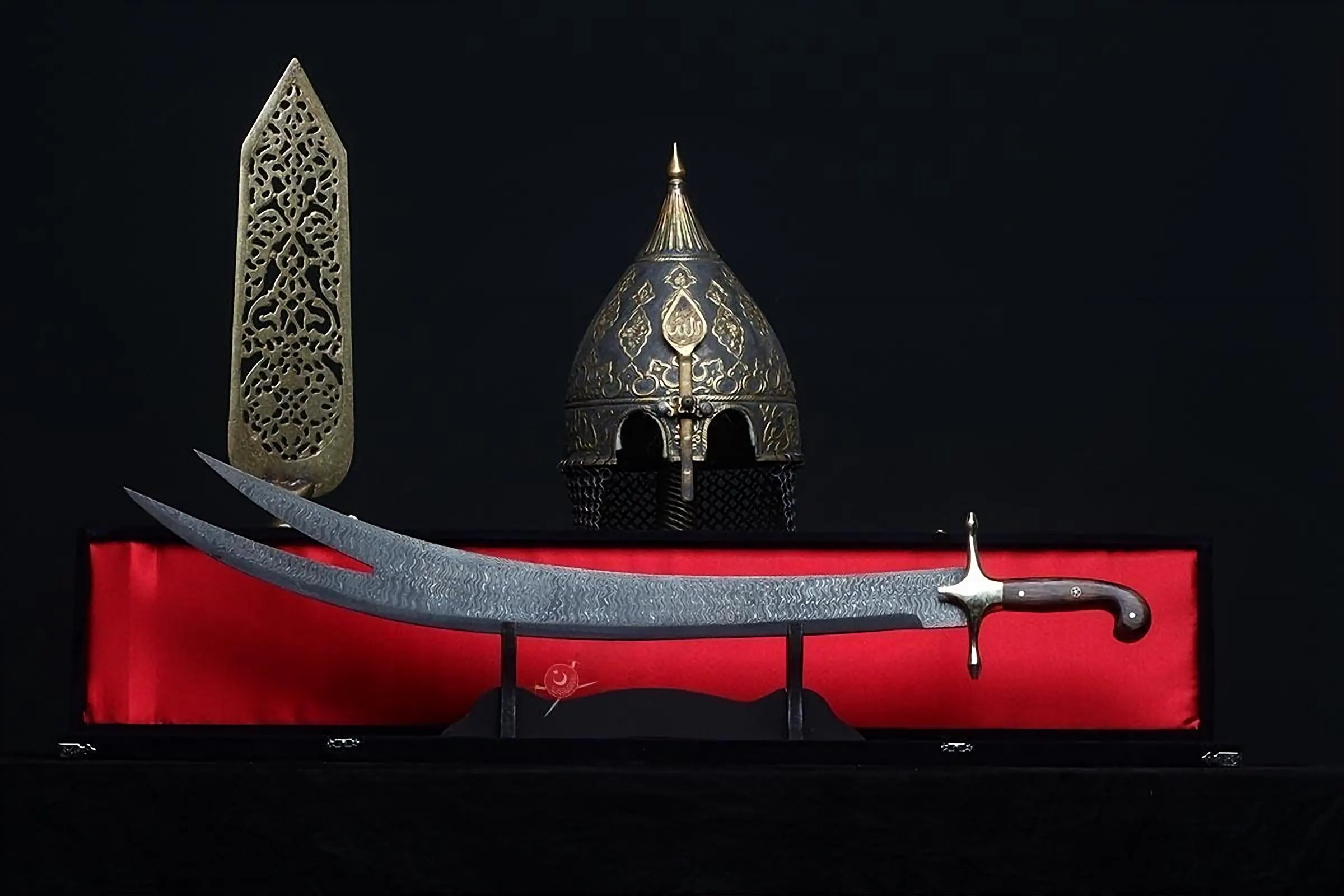 A curved double-pointed sword with a dark textured blade and a wooden handle, displayed horizontally on a stand. Behind it sits a pointed metal helmet with gold-coloured engravings and a tall, ornate, flat metal decorative piece.