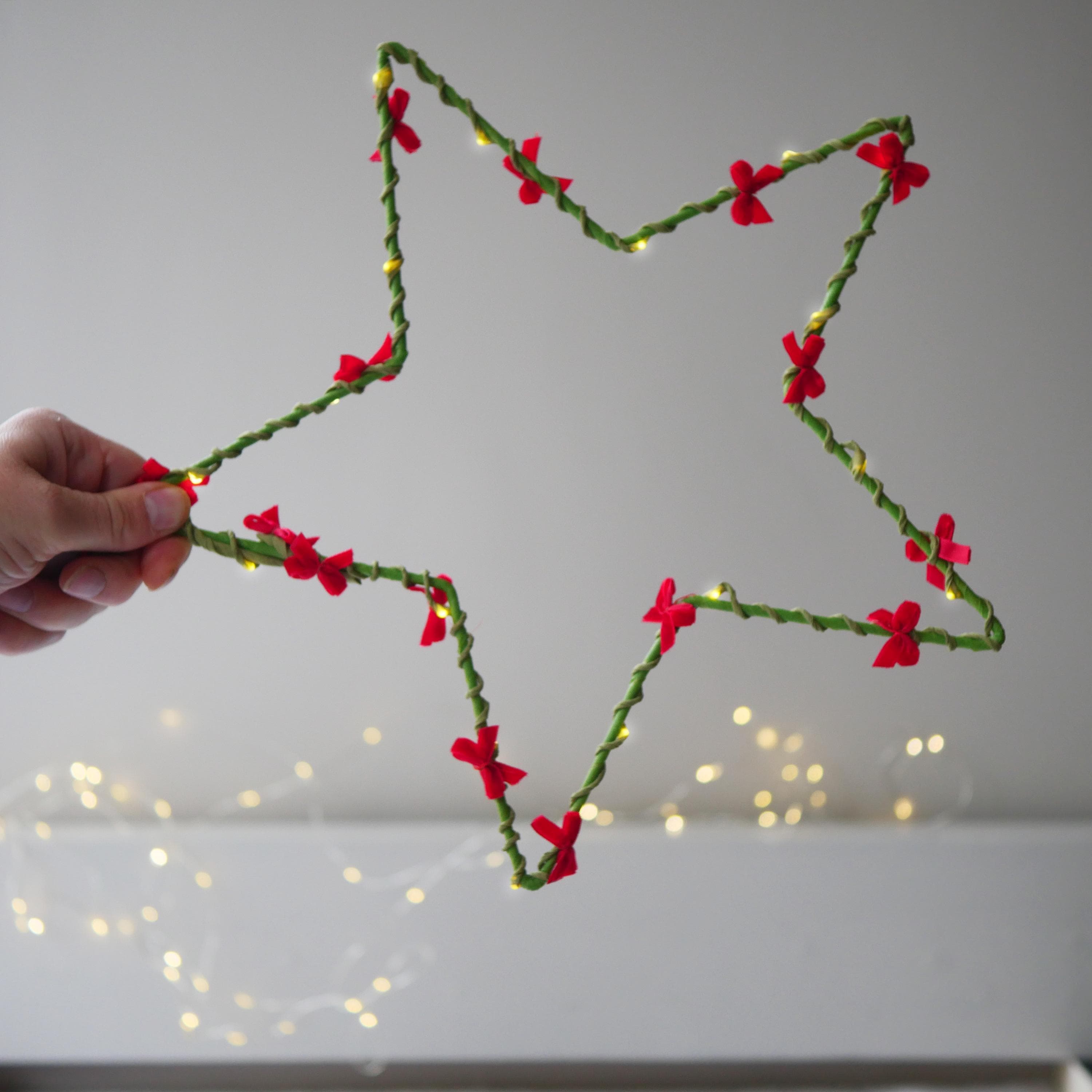 May include: A green star-shaped decoration adorned with red bows and small yellow lights. The star is held by a hand against a neutral background. A festive holiday decoration.