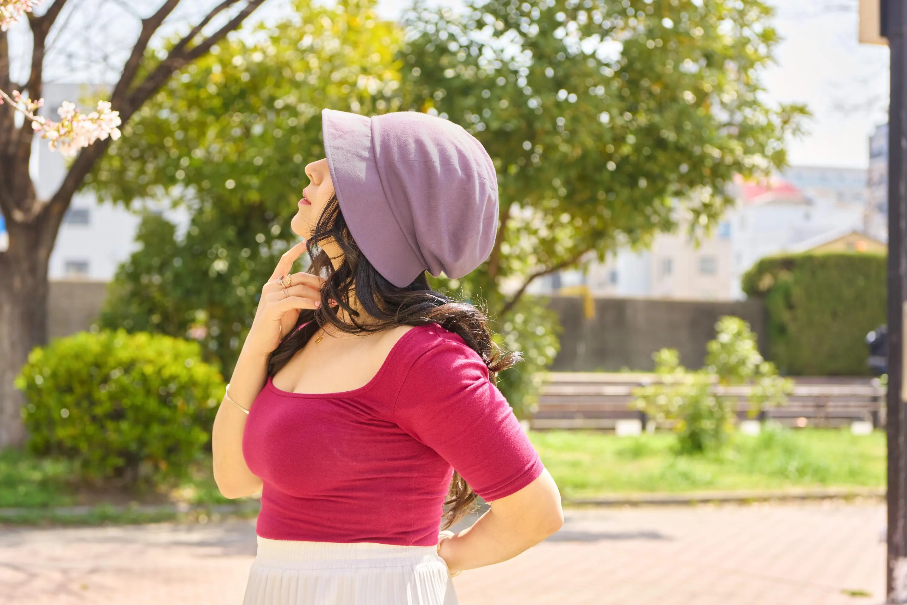May include: A woman wearing a purple hat and a red top, standing outdoors with a green lawn and trees in the background.